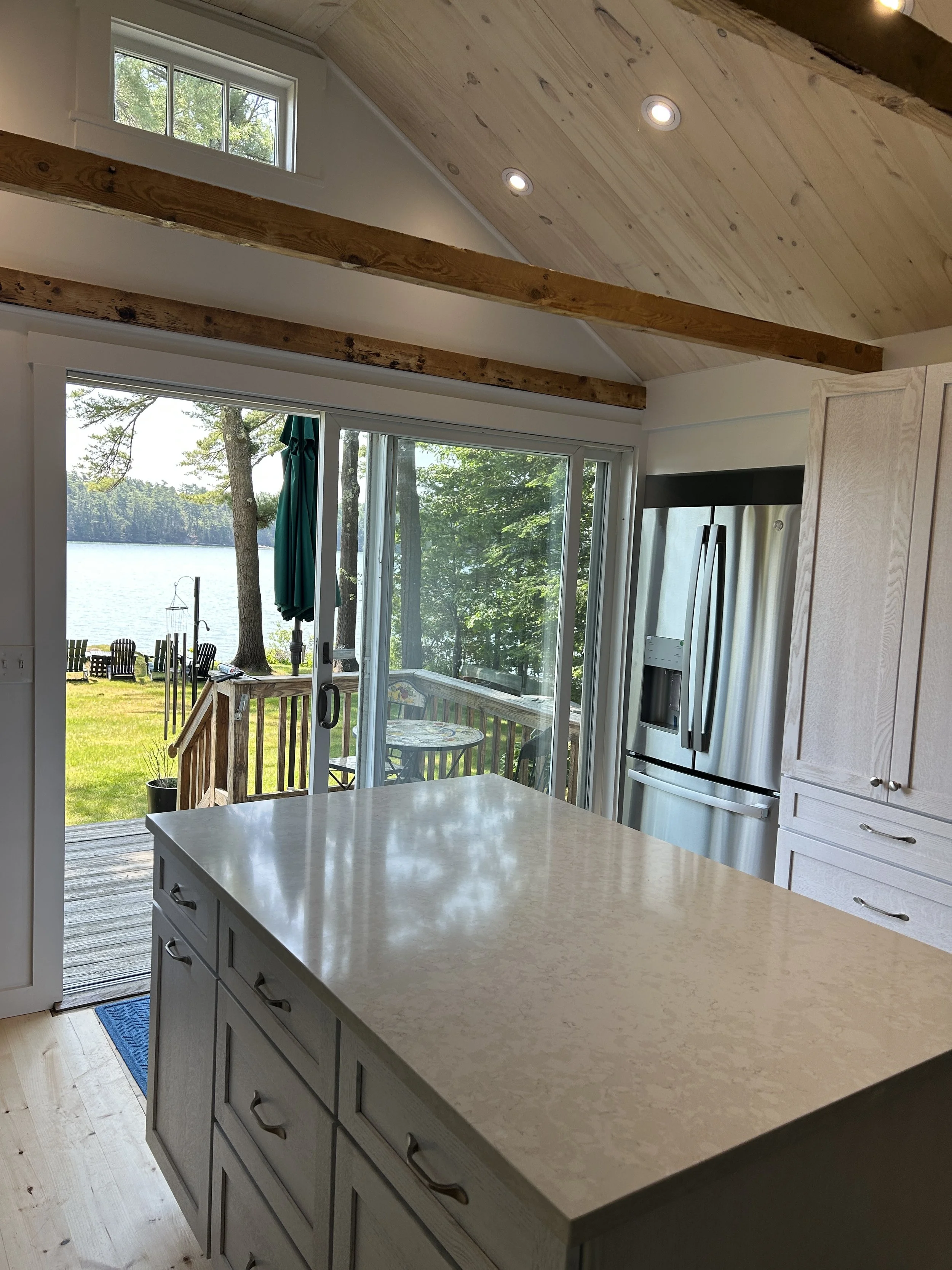 A view from inside a kitchen looking out through sliding glass doors to a lakeside scene with trees, chairs, and an umbrella on a deck.