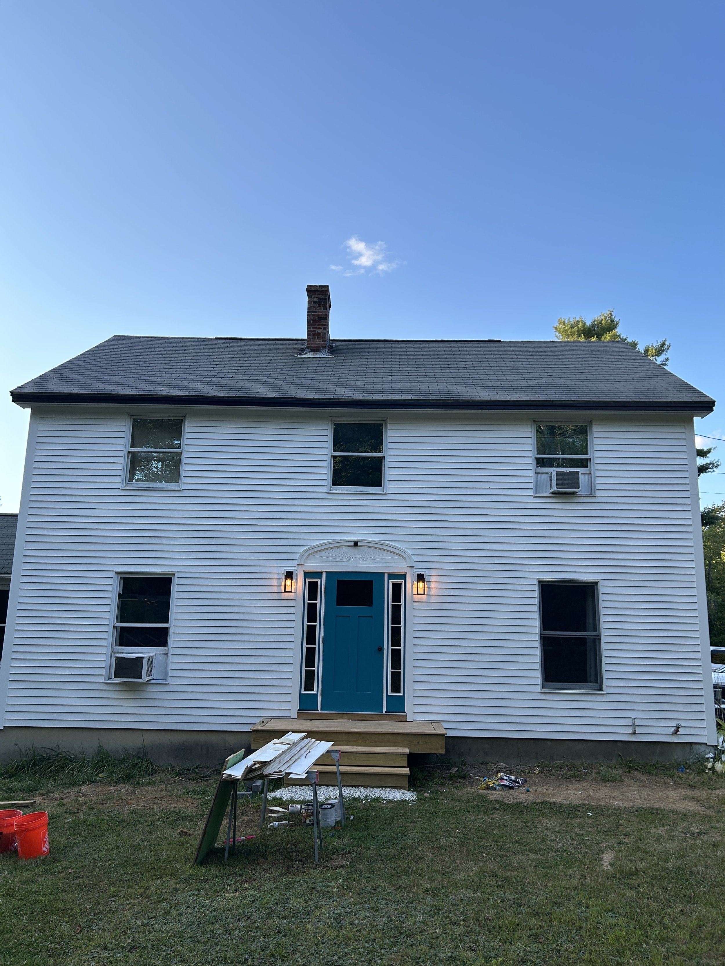 The back of a two-story white house with green grass in front, under a clear blue sky with a few clouds. The house has a blue front door with sidelights and a transom window, and four windows on the second and first floors. There is an air conditioni