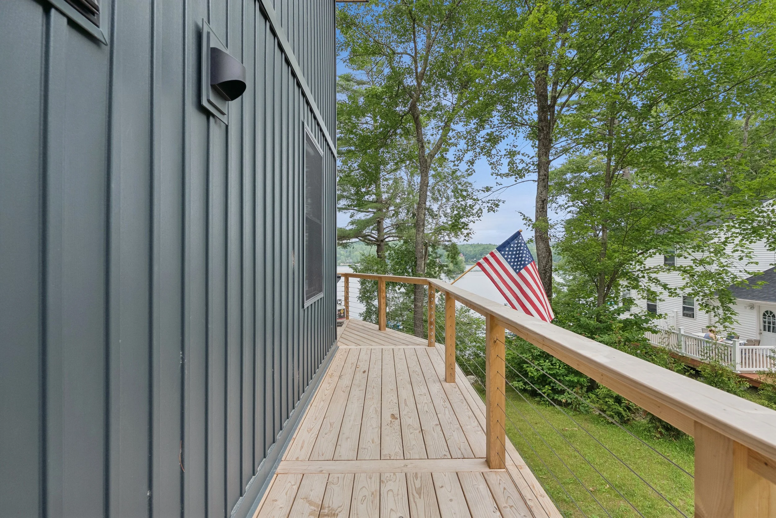 A wooden balcony with a metal railing overlooking green trees and houses, with an American flag hanging from the railing.