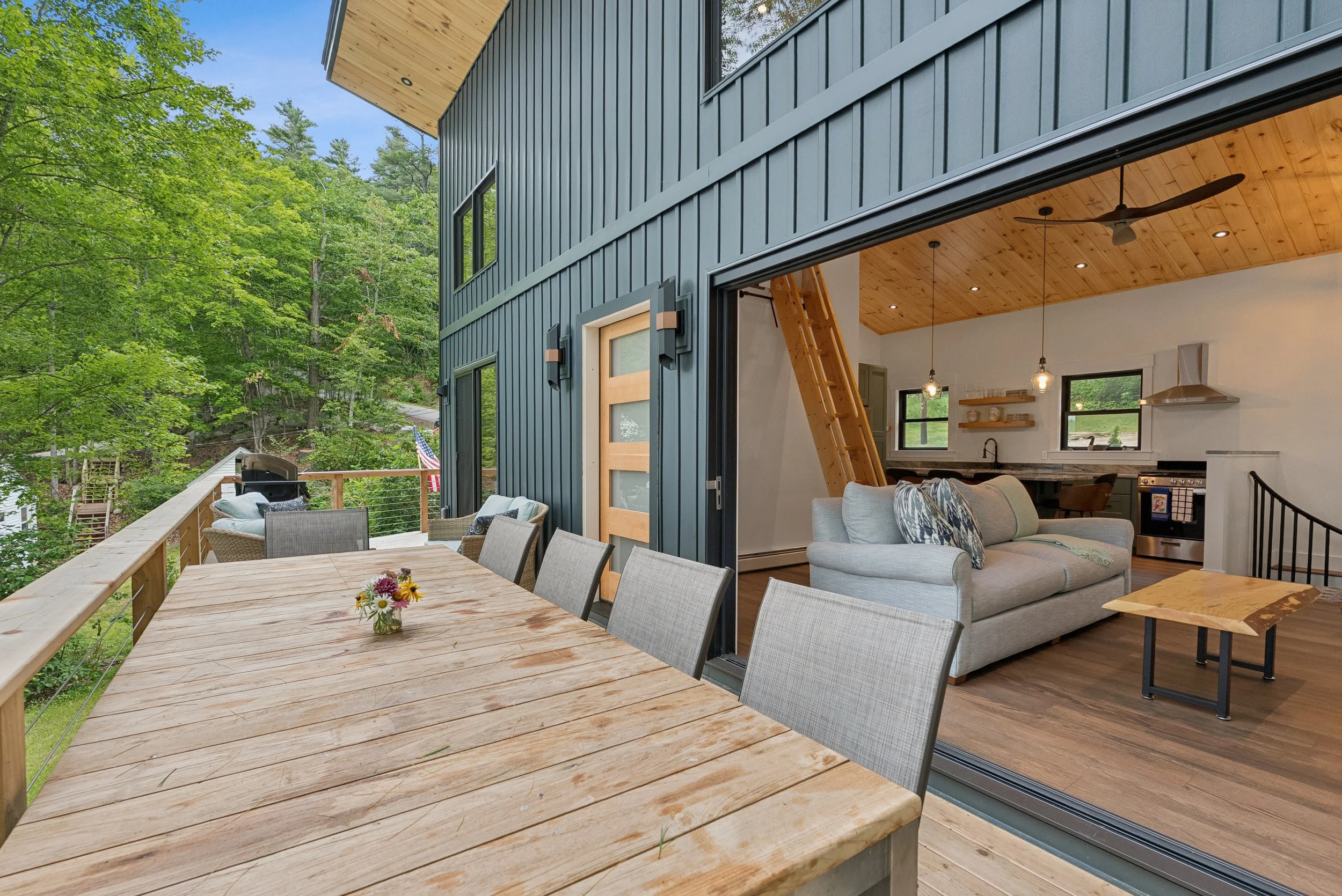 Deck with outdoor dining table and chairs overlooking a lush green forest, with the interior of a modern house visible through sliding glass doors.