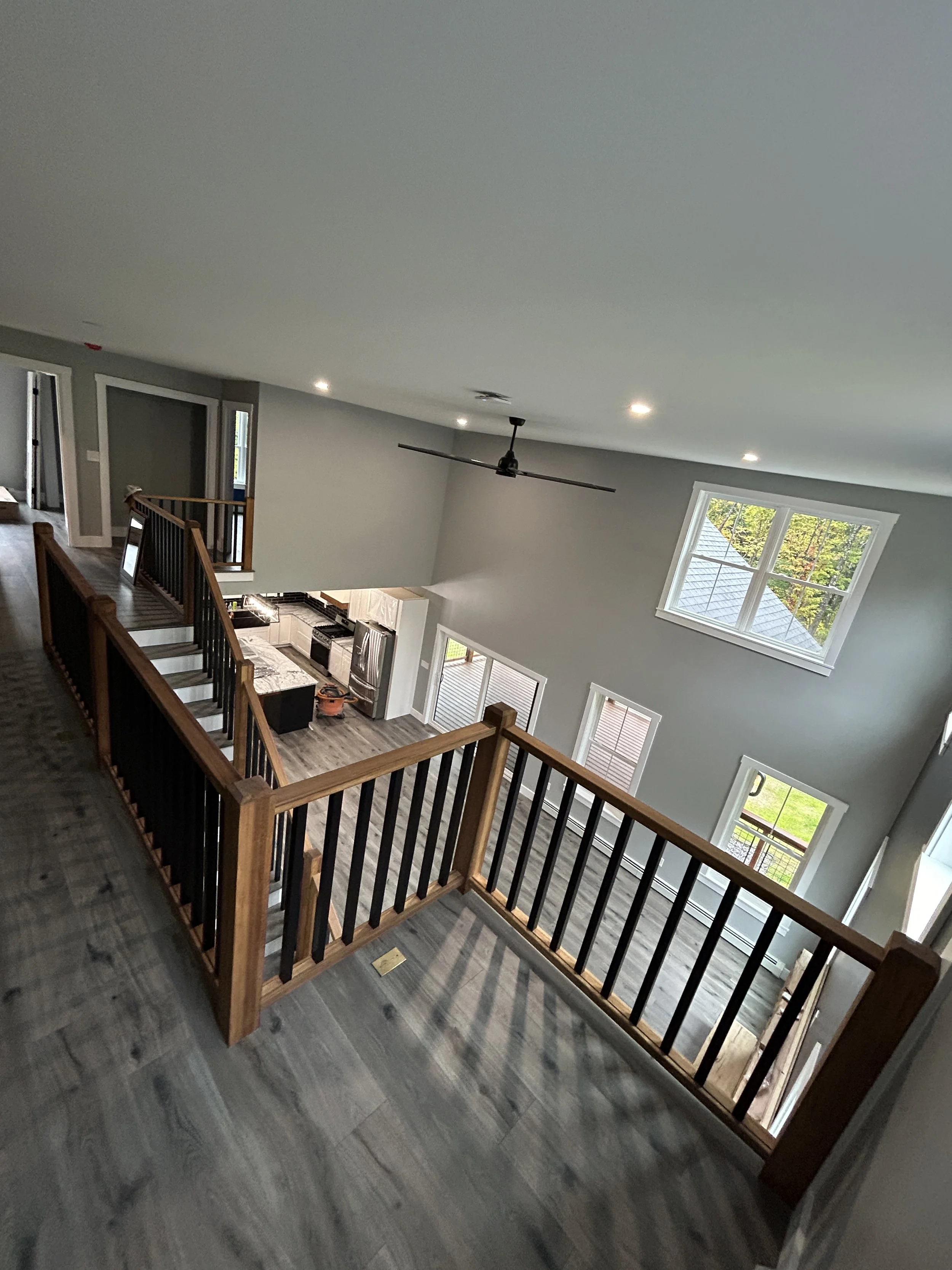 Interior view of a two-story modern home with a spacious open floor plan, featuring large windows, gray walls, hardwood flooring, and a black ceiling fan with lights. The upper level has a wooden railing overlooking the lower level, which includes a 
