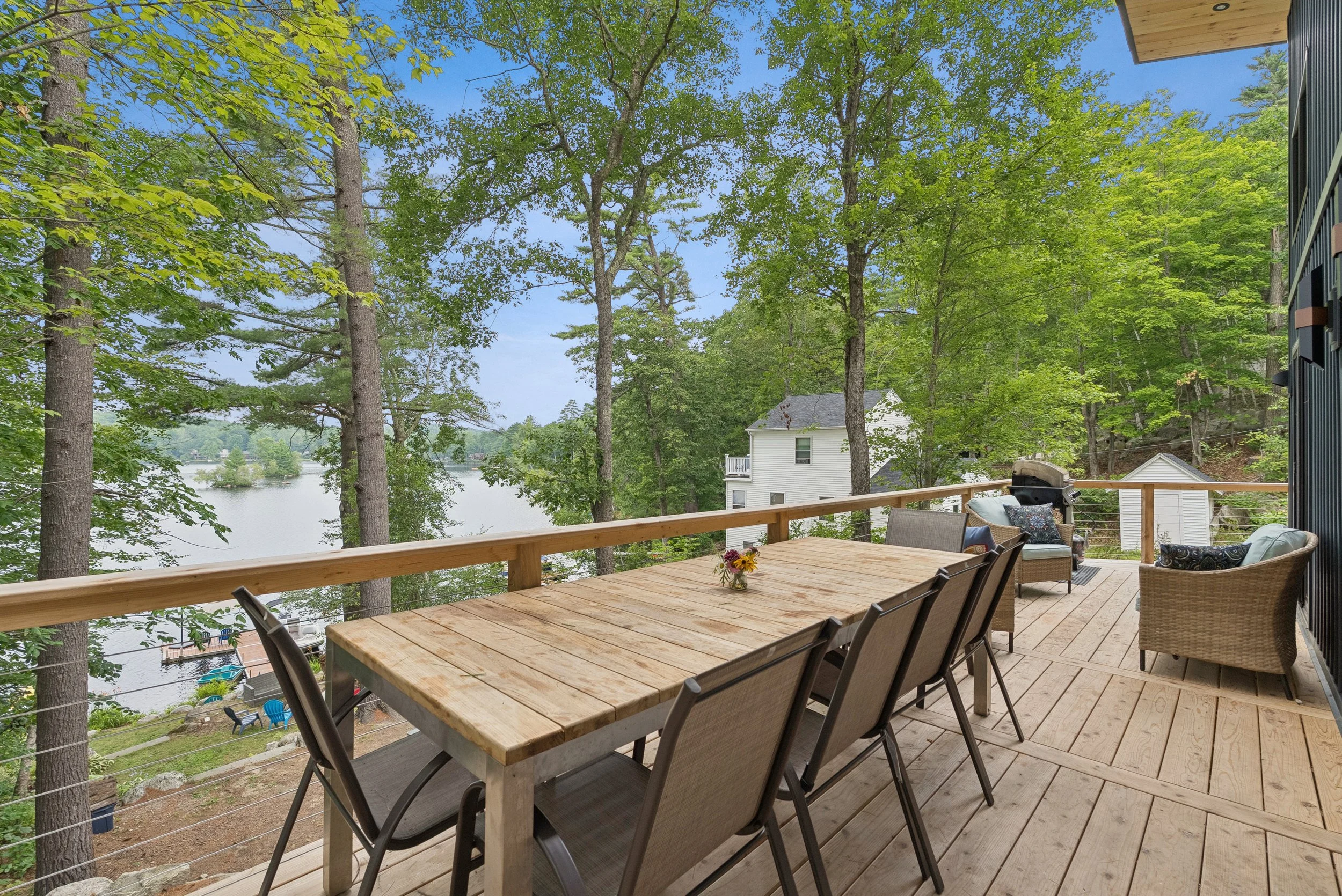 A wooden deck outdoor patio overlooking a forested area with tall green trees and a distant house near a lake, decorated with outdoor furniture including a table with a vase of flowers, chairs, and a grill.