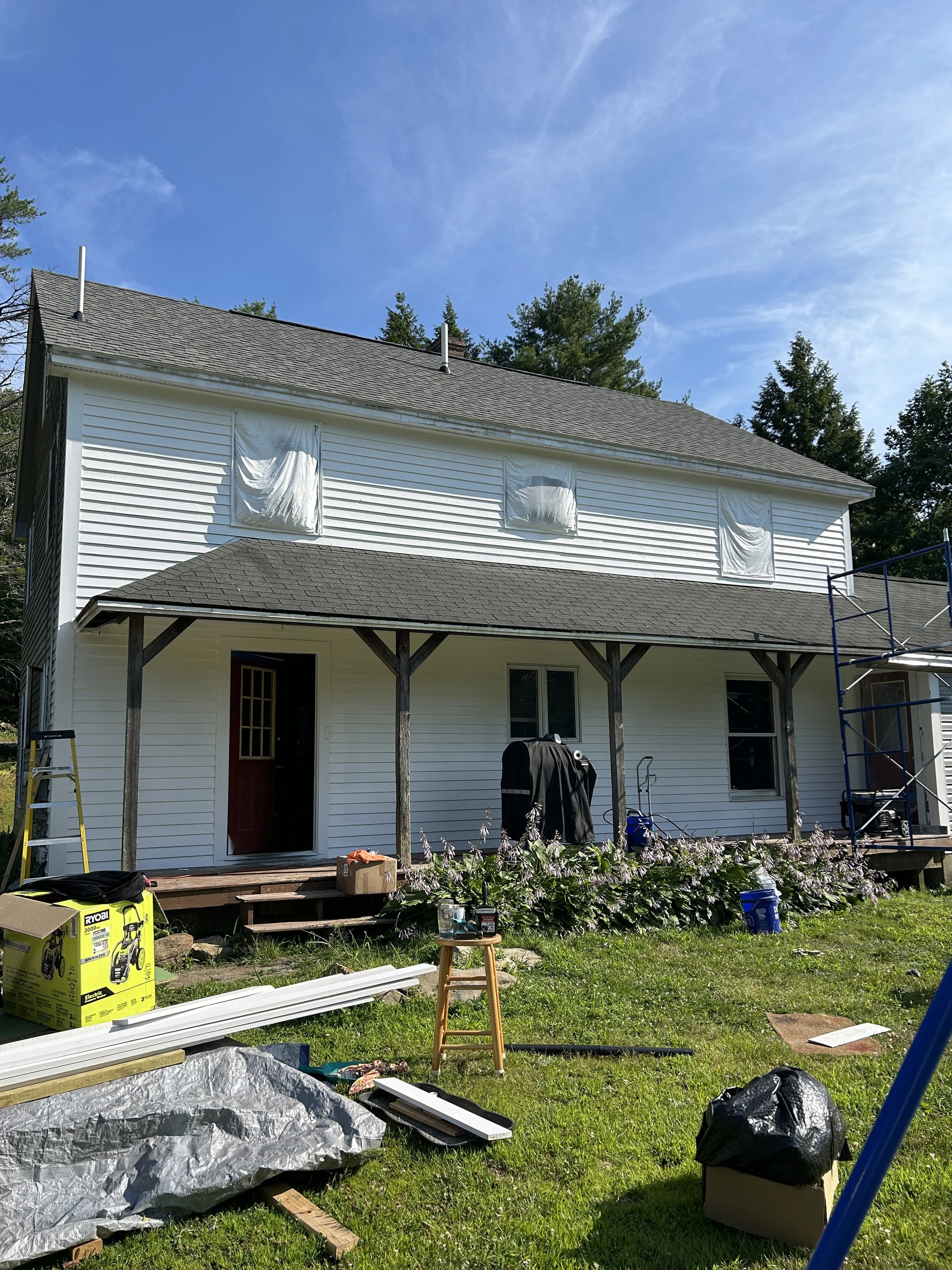 Back view of a house under renovation, with white siding, a covered porch, and construction materials and tools surrounding the yard.