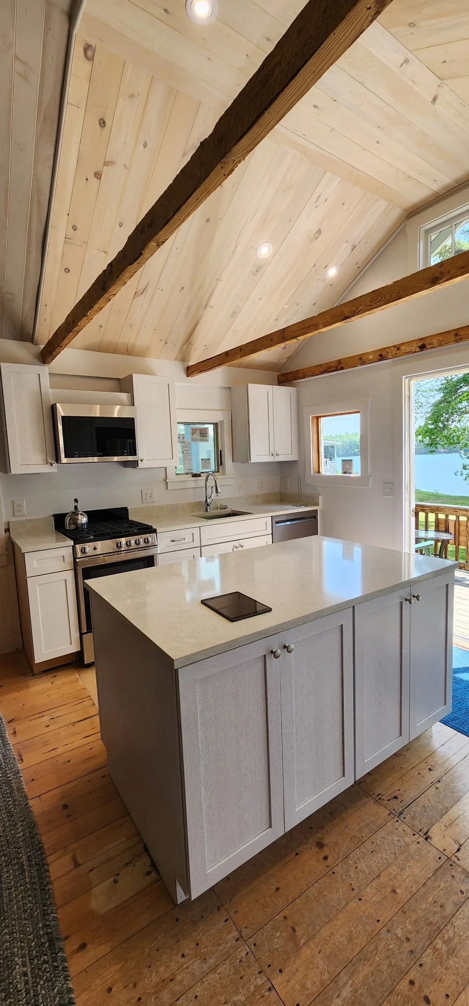 A bright kitchen with a sloped light wood ceiling and exposed wooden beams. It features white cabinetry, a stove, microwave, and an island with a smooth countertop. There are windows and a door leading to an outdoor deck with a table and chairs.