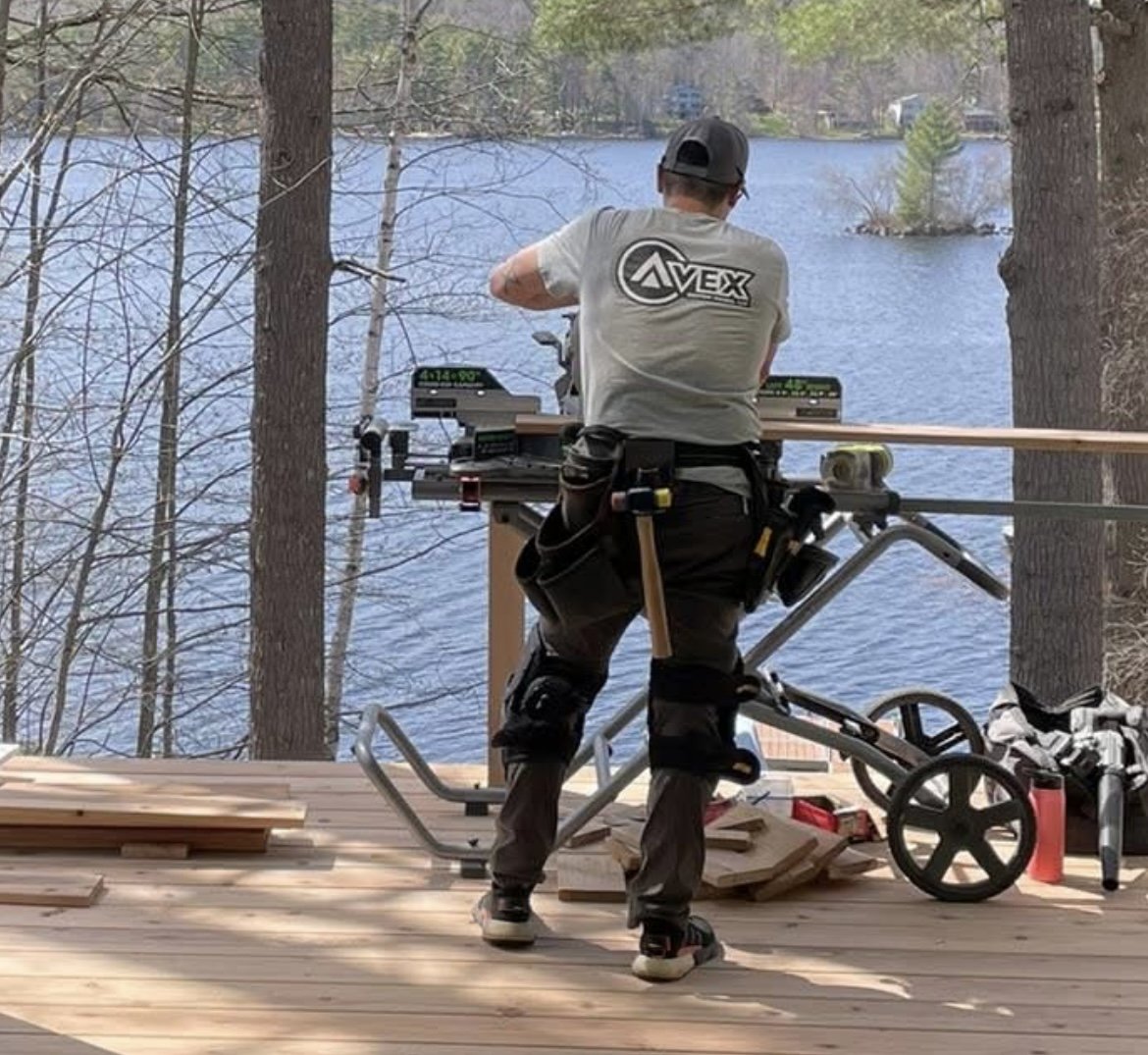 Avex Custom Homes owner Andrew is working on a carpentry project outdoors on a wooden deck by a lake, with trees and water in the background.