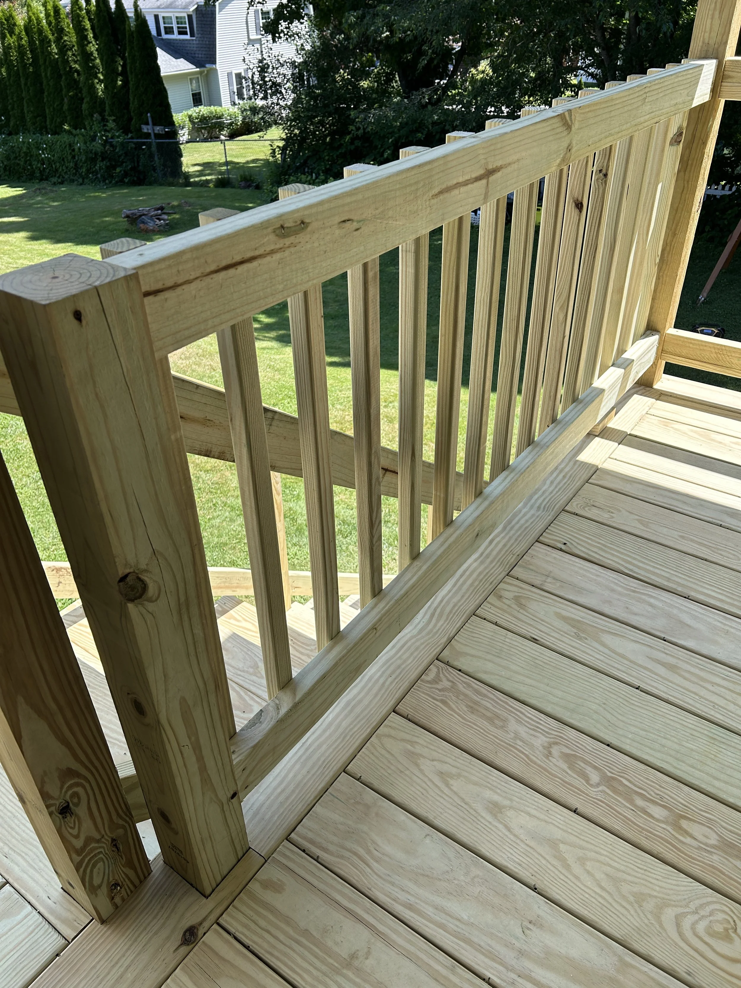 Wooden outdoor deck railing with vertical slats, view of a green backyard with trees, shrubs, and a neighboring house in the background.