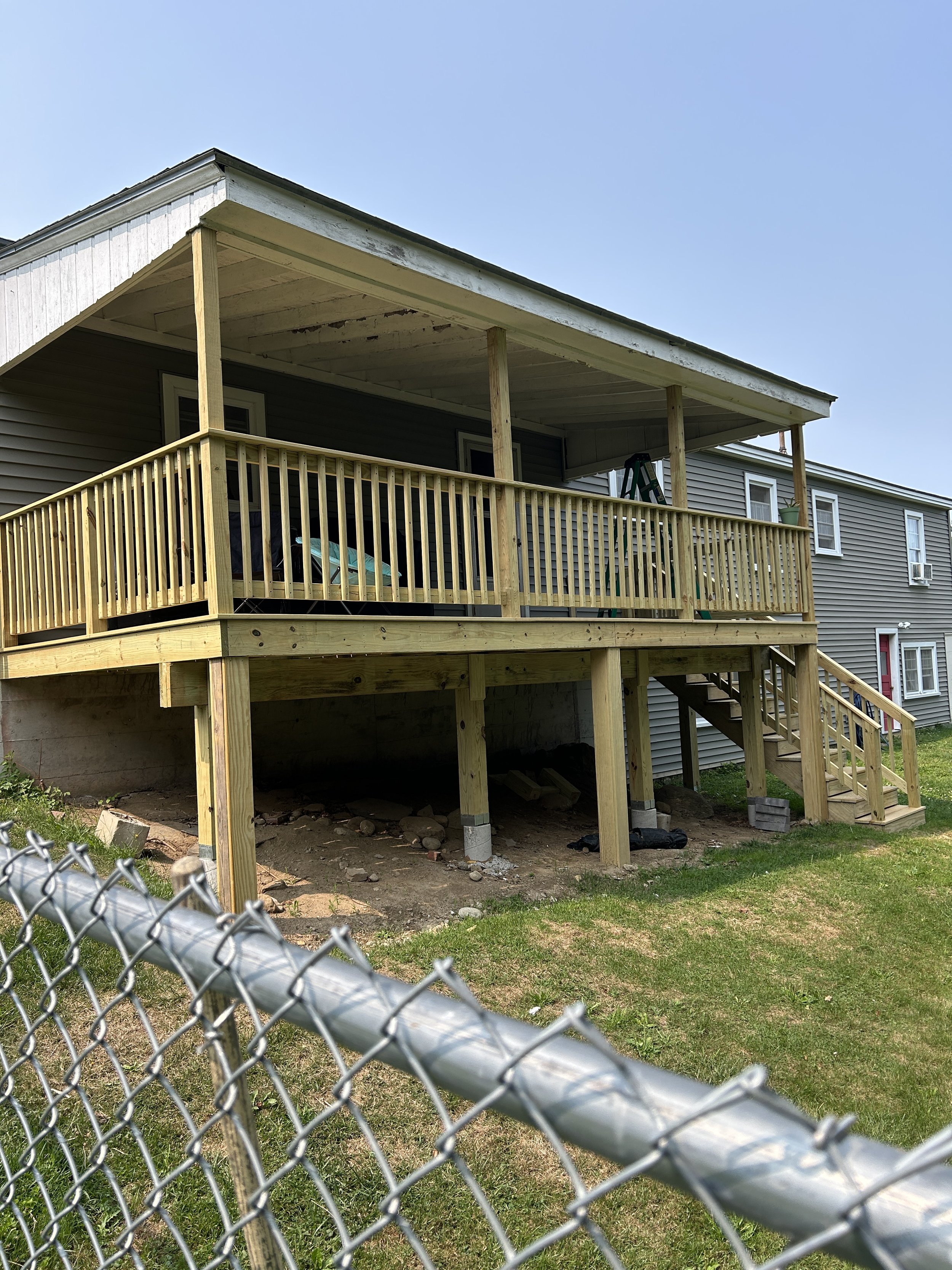 A house with a wooden deck under construction, featuring a staircase, railing, and support beams, with some construction materials underneath and a fence in the foreground.