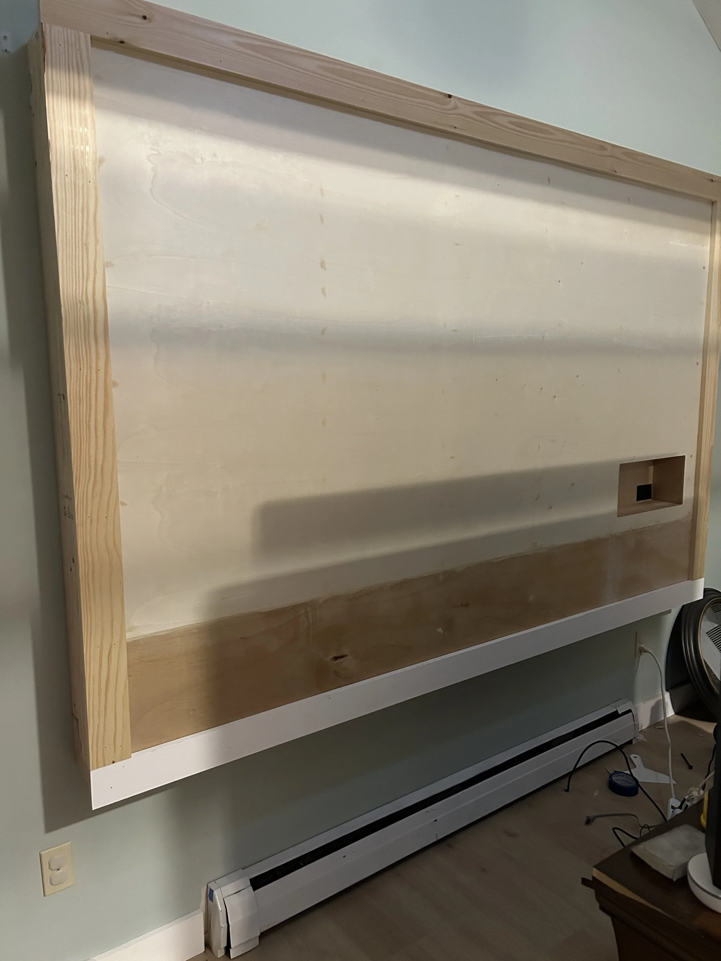 The image shows a wooden tv frame structure, mounted on a green wall, with an electrical outlet and heater at the bottom.