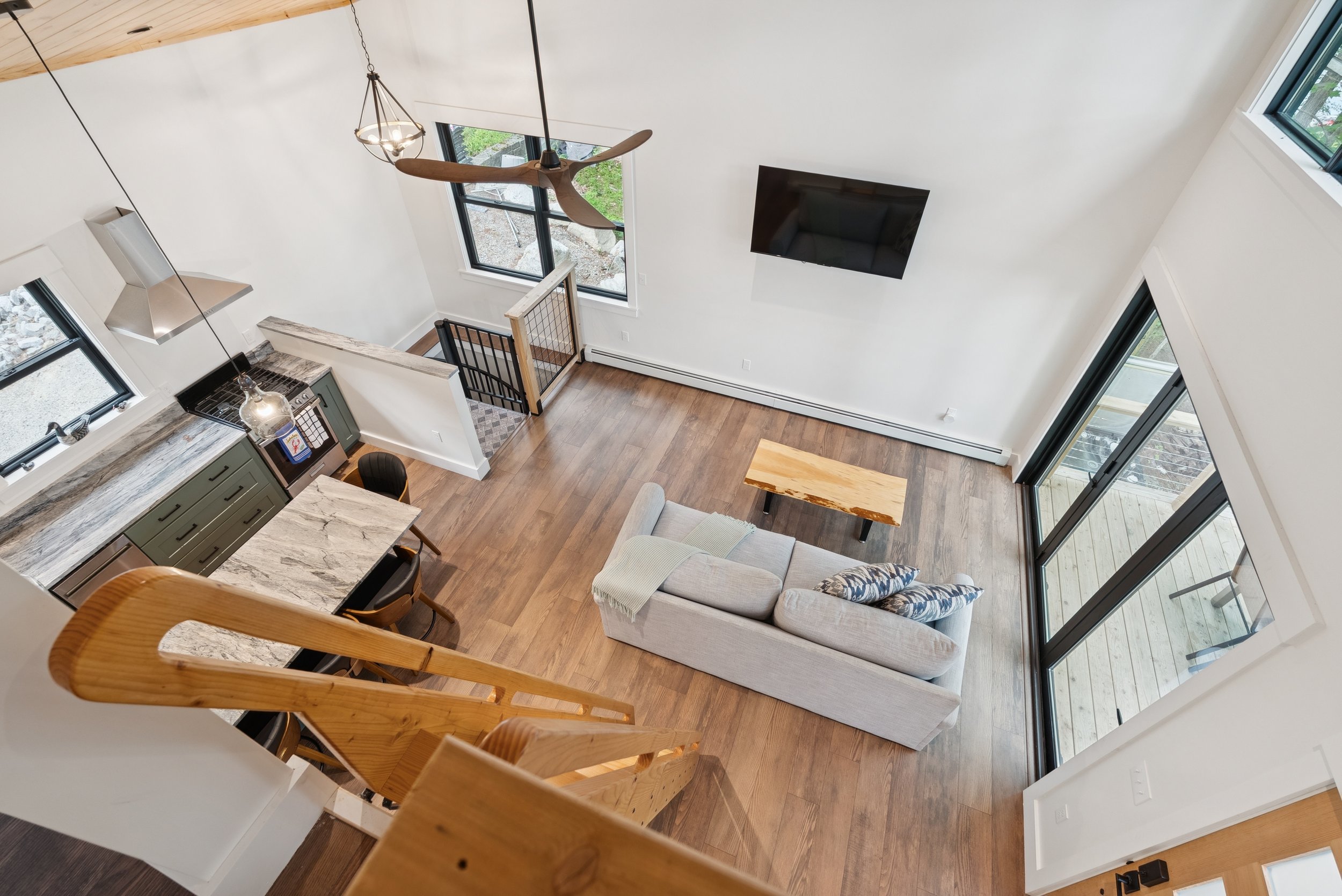 An aerial view of a modern living room with a cozy gray sofa, a wooden coffee table, and a flat-screen TV on white walls. The space features large windows offering natural light and a view of outdoor greenery, with a staircase with wooden railing in 