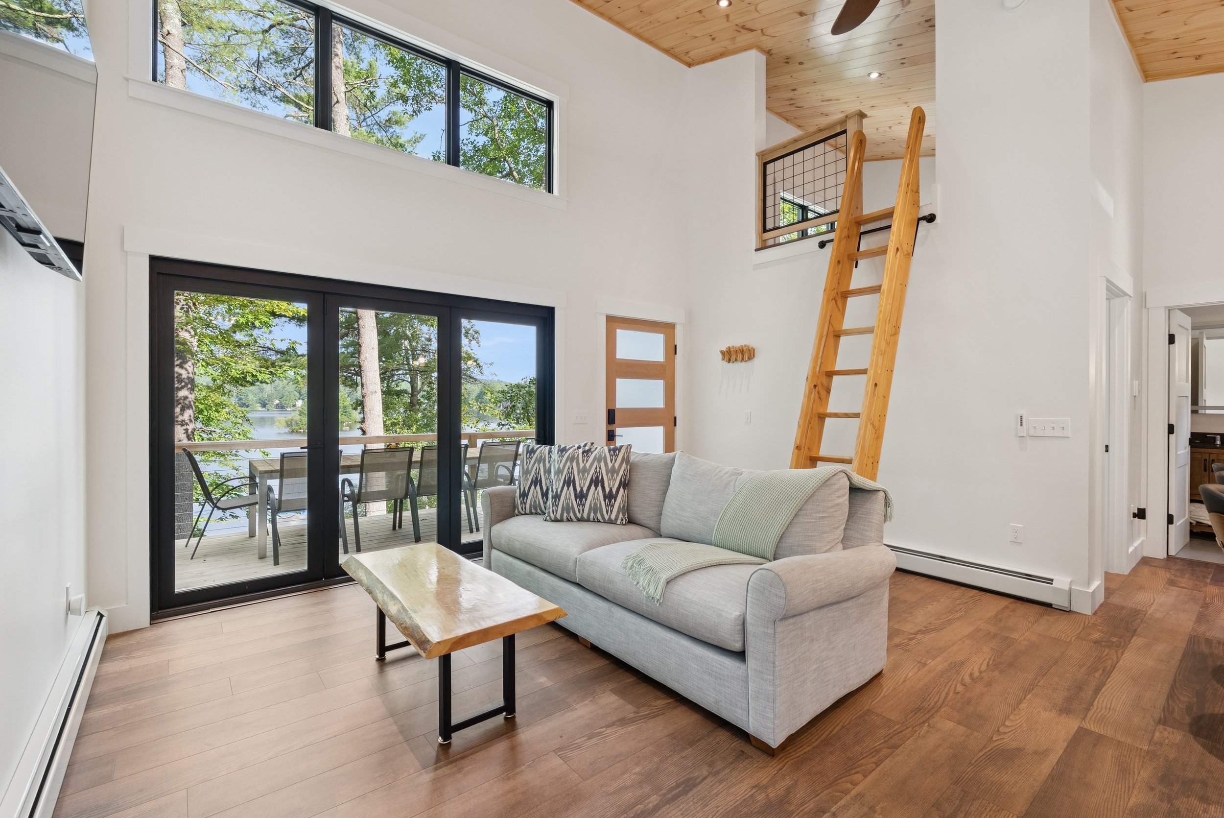 Living room with high ceiling, large windows showing trees and lake view, sliding glass door leading to balcony with outdoor seating, light-colored sofa with patterned pillow, wooden coffee table, sky-colored throw blanket, wood flooring, and wooden 