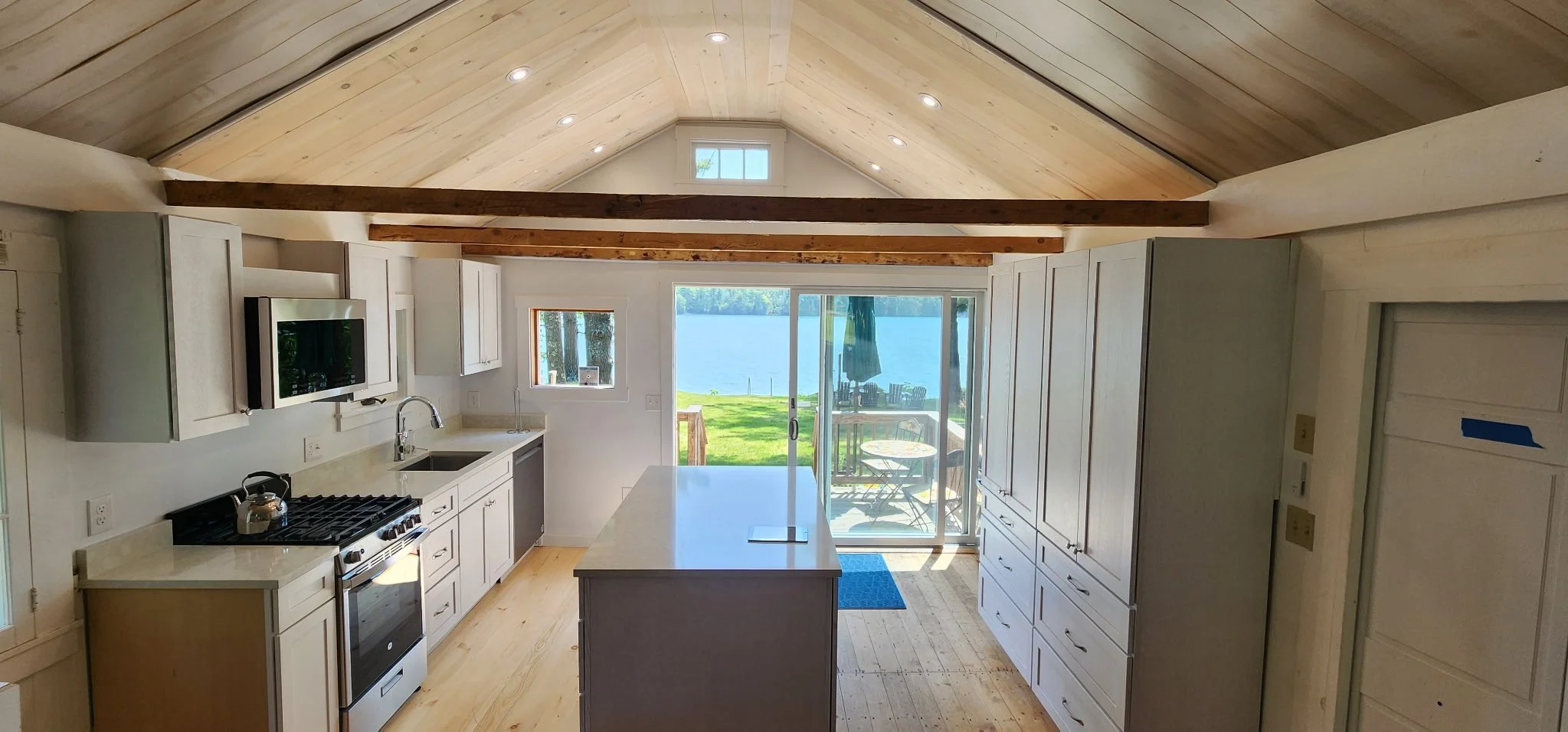 Kitchen with white cabinets, a stove, a sink, and a large window and sliding glass door showing an outdoor scene with a lake, trees, and a patio table.