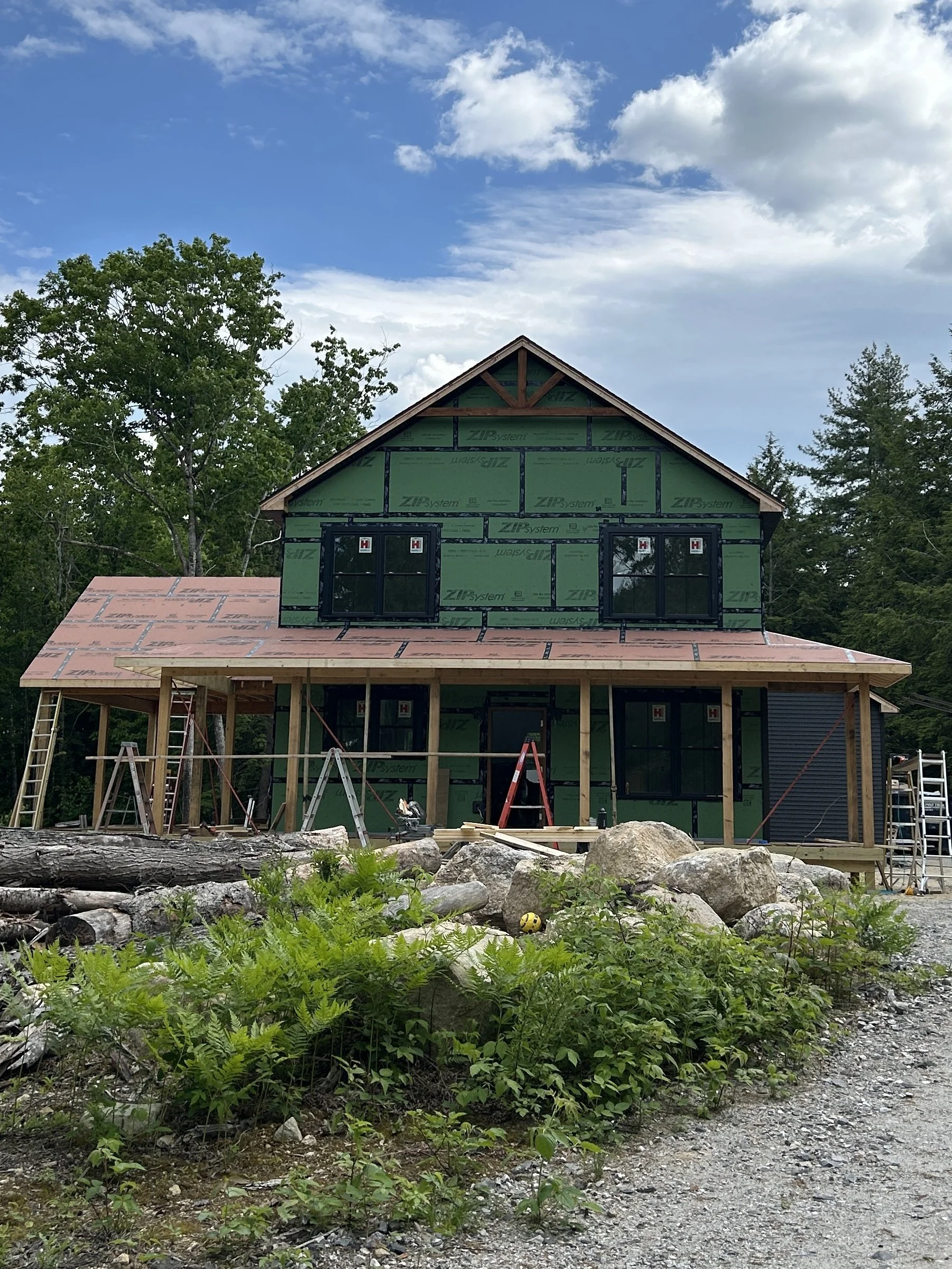 A house under construction with a partially completed roof and green sheathing. Construction tools and ladders are visible, and the house is surrounded by rocks, plants, and trees.
