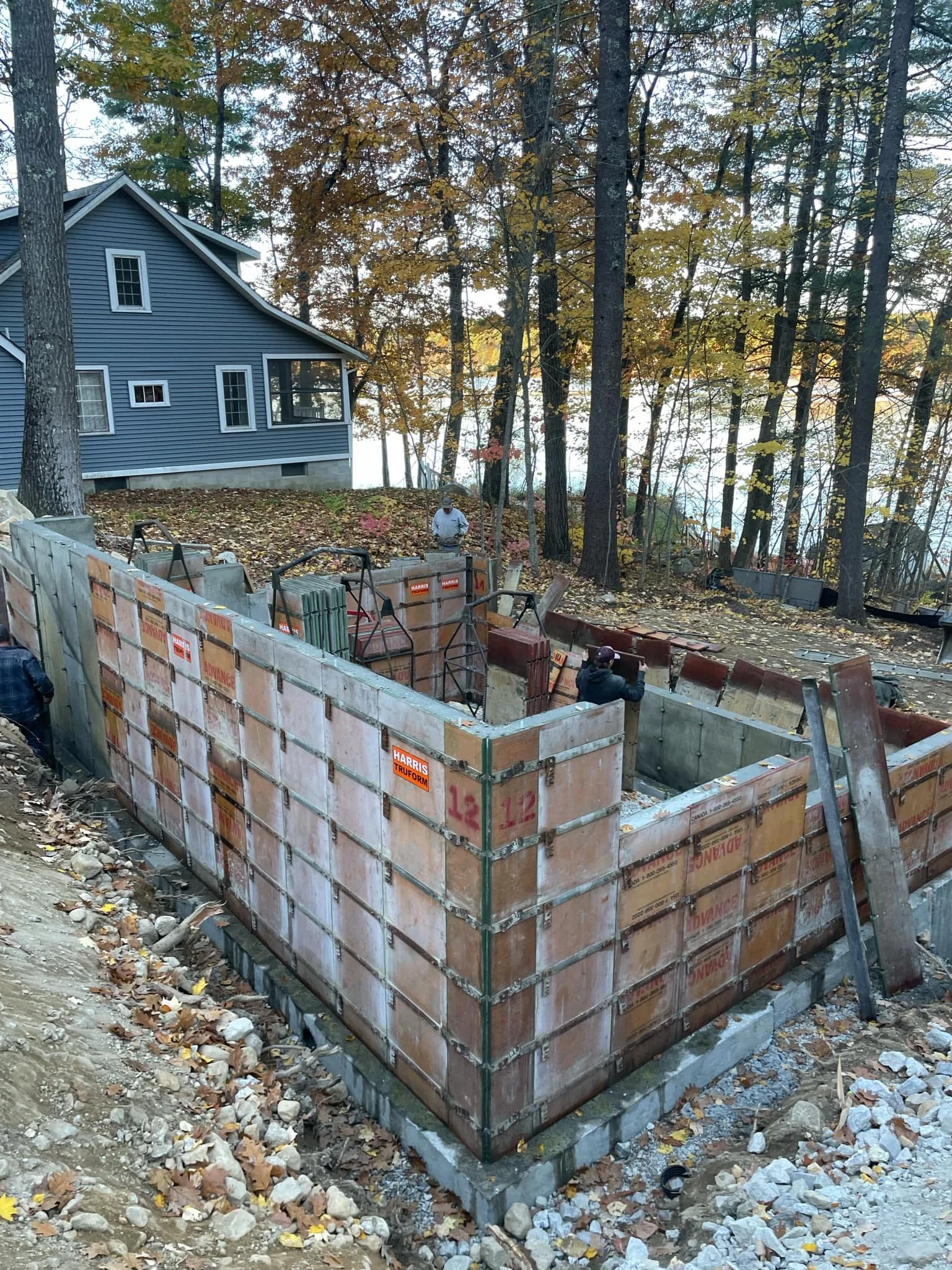 Construction site with a concrete foundation being built, surrounded by trees with fall foliage and a blue house in the background.