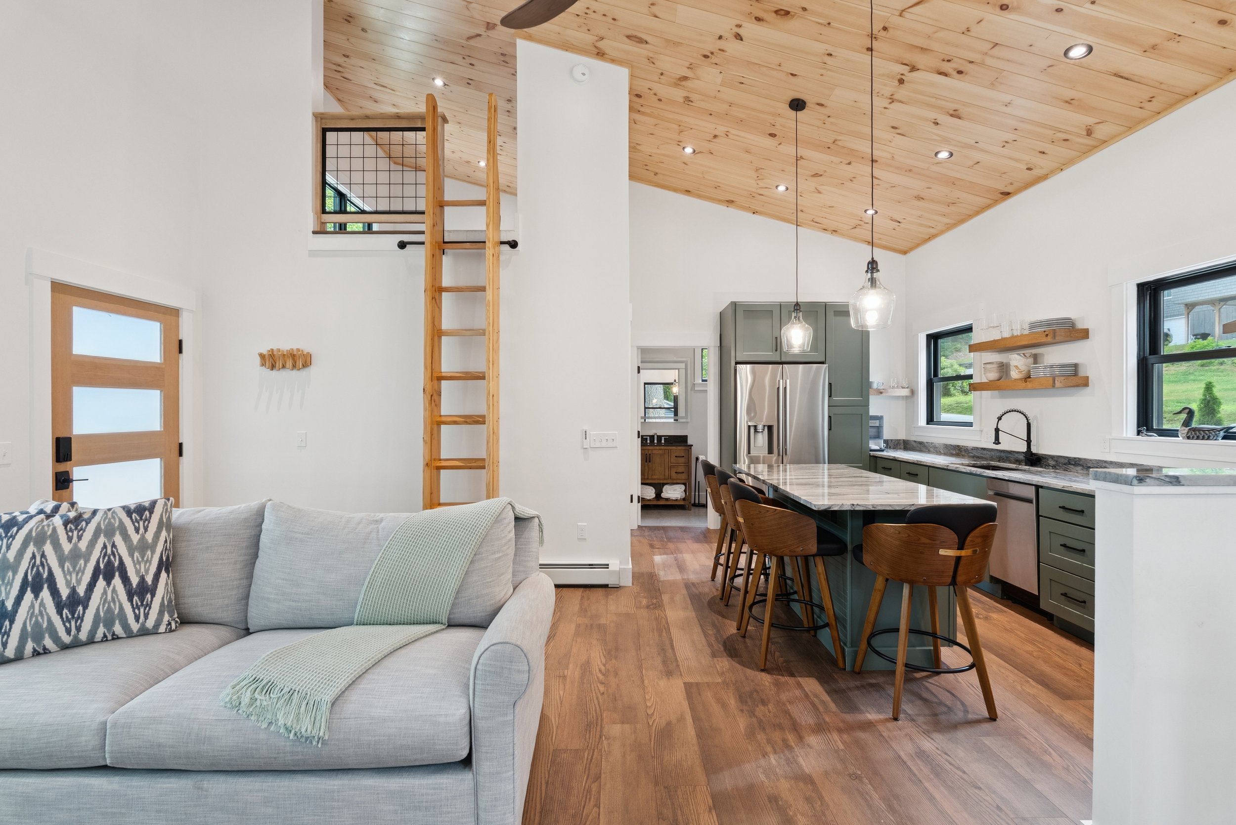 Open-concept living room and kitchen with wooden vaulted ceiling, white walls, and hardwood floors. The living room has a light-colored sofa with patterned pillows, and the kitchen features green cabinets, a marble island, open wooden shelves with di