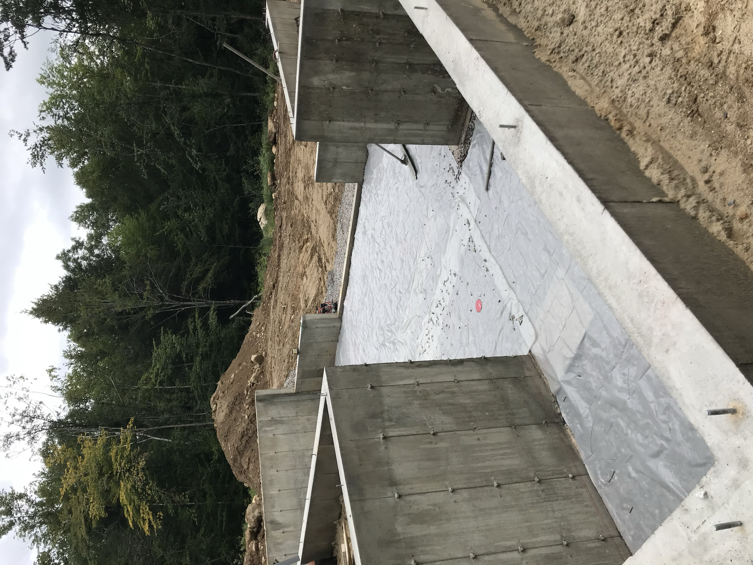 Wooden fence with concrete foundation, construction site with white covering on ground, and trees and sky in the background.