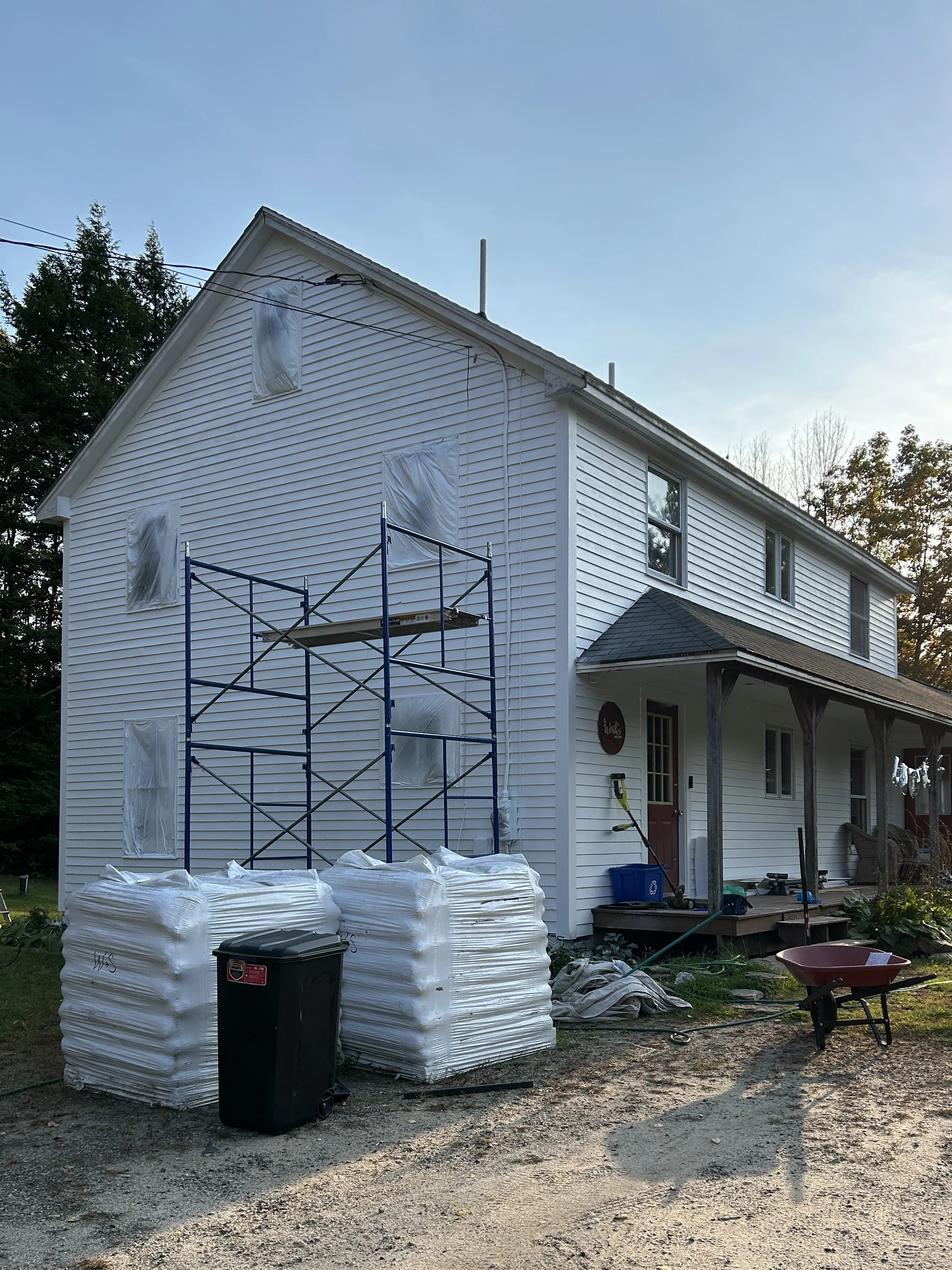 Side view of a two-story house with white siding, scaffolding outside, covered windows, and construction materials and tools in the yard.
