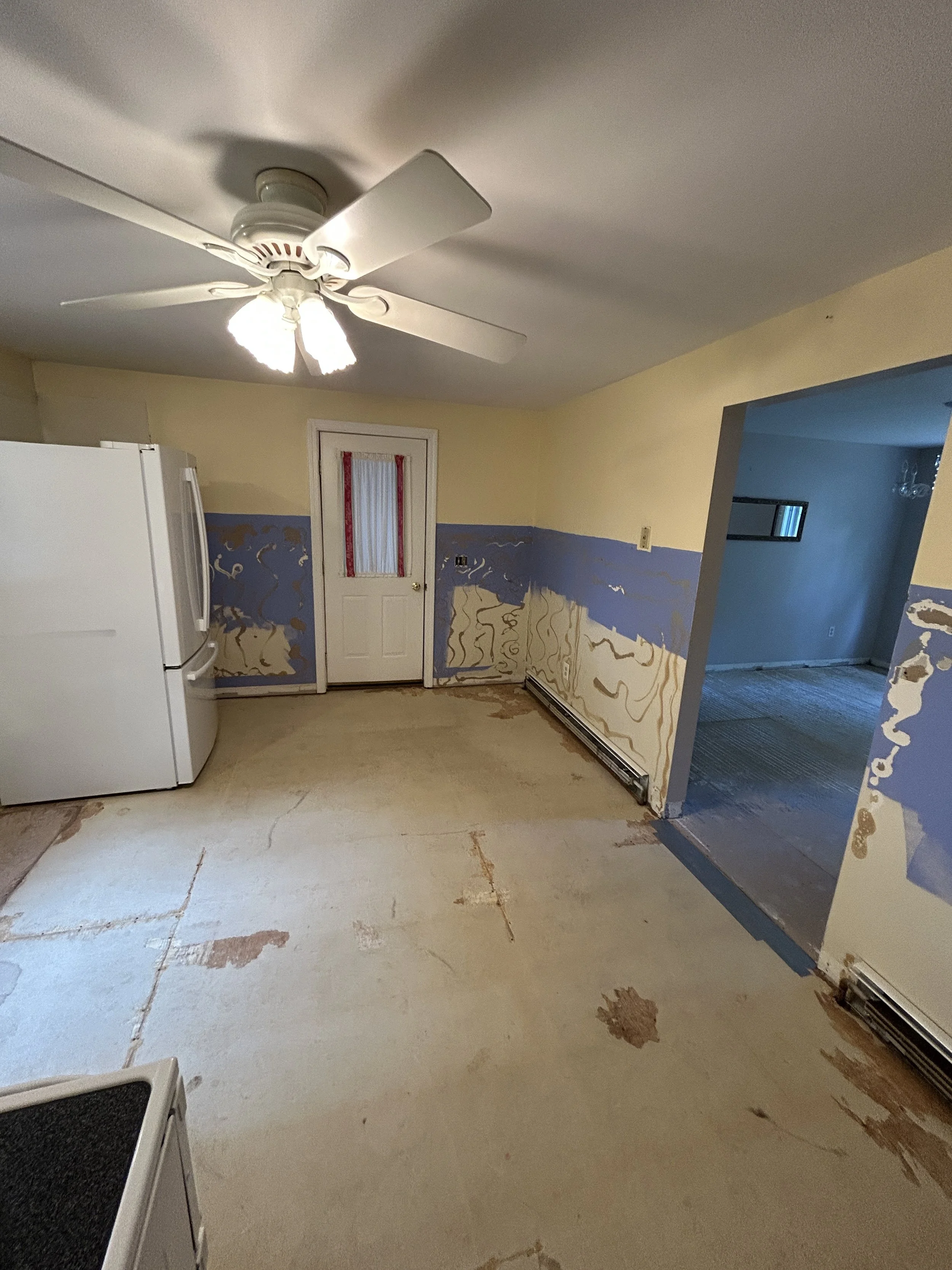 A room with a ceiling fan and light fixture, partially renovated with visible patches of drywall and peeling paint, an open doorway leading to a darker room, and a white refrigerator in the corner.