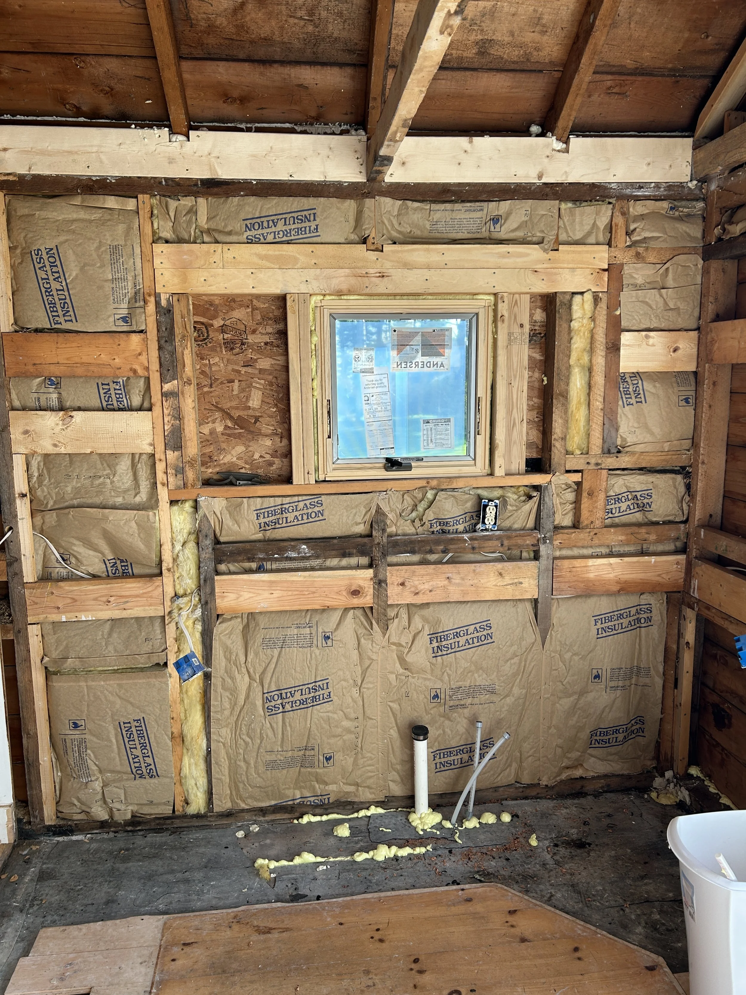 The interior of a building under construction with exposed wooden framing, insulation, a small window, and plumbing pipes.