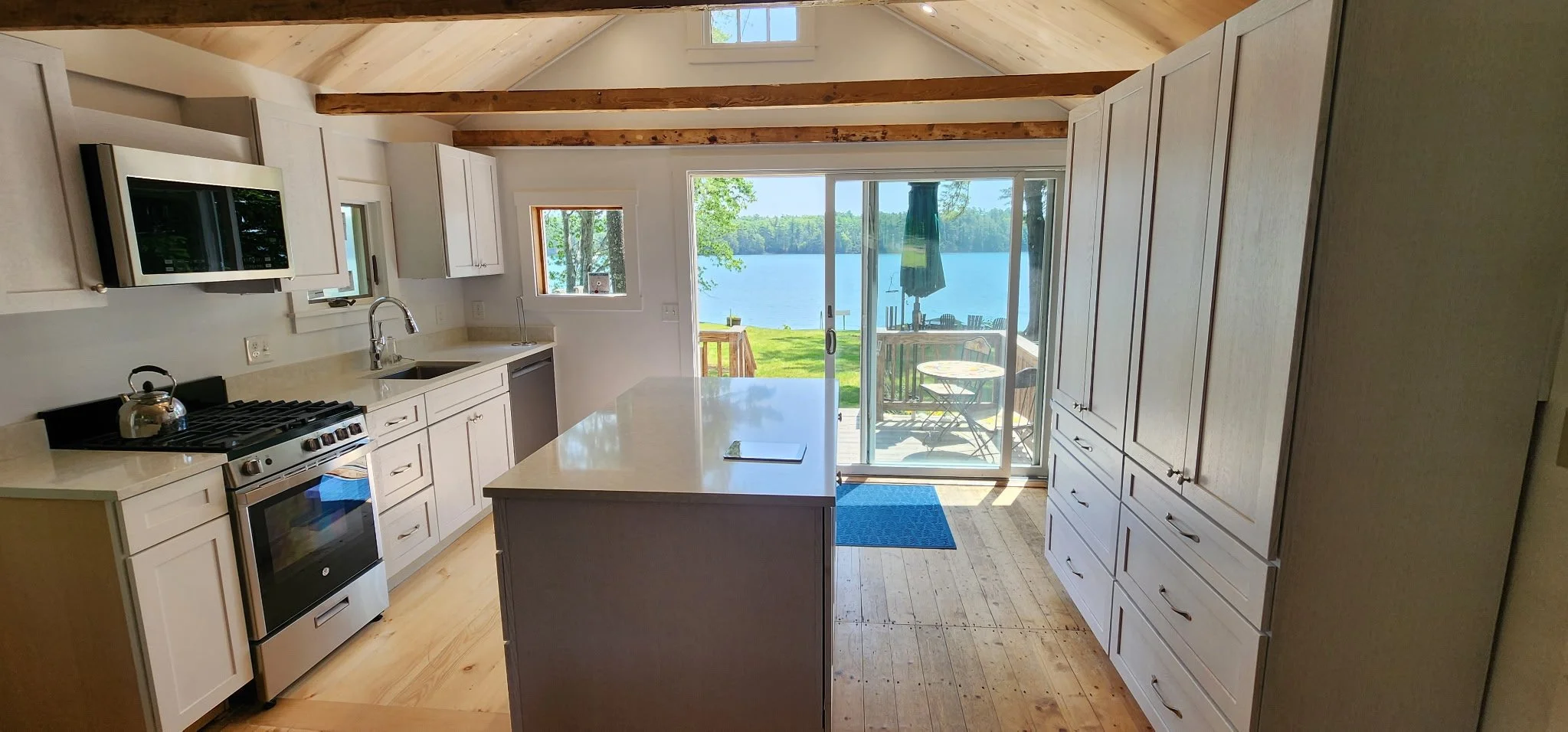 Kitchen with white cabinets, beige countertops, a stove with a kettle, a microwave, and a sliding glass door leading to a deck with outdoor furniture. Large windows display a lake view with green trees.