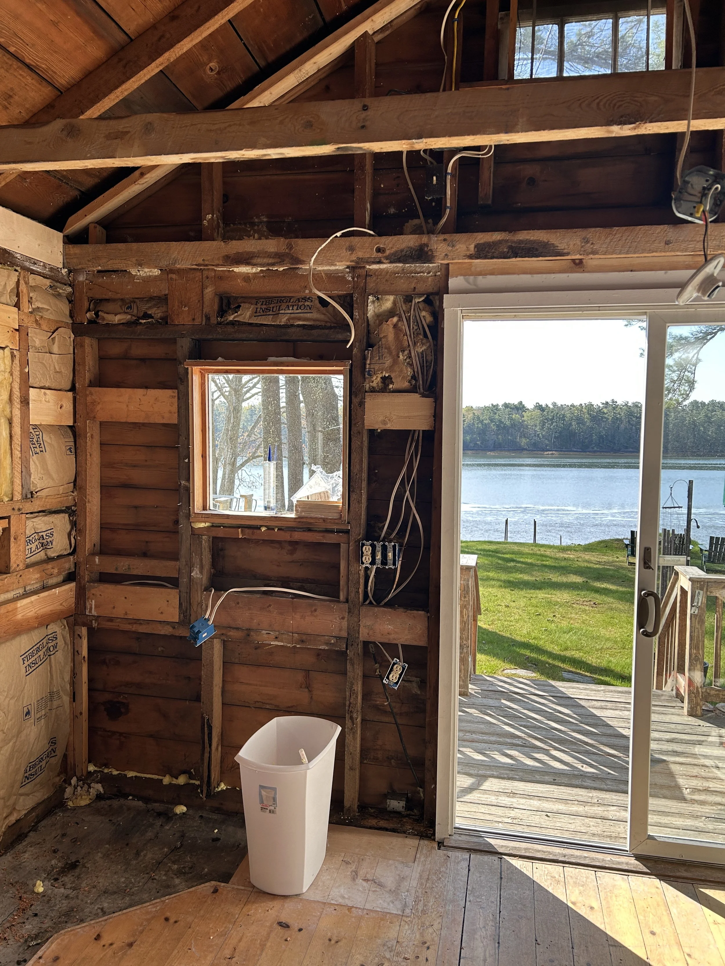 A partially remodeled room with exposed wooden wall studs, electrical wiring, and insulation. A sliding glass door opens to a view of a lake with trees across the water. There is a white trash can on the floor, and an opening in the wall shows constr