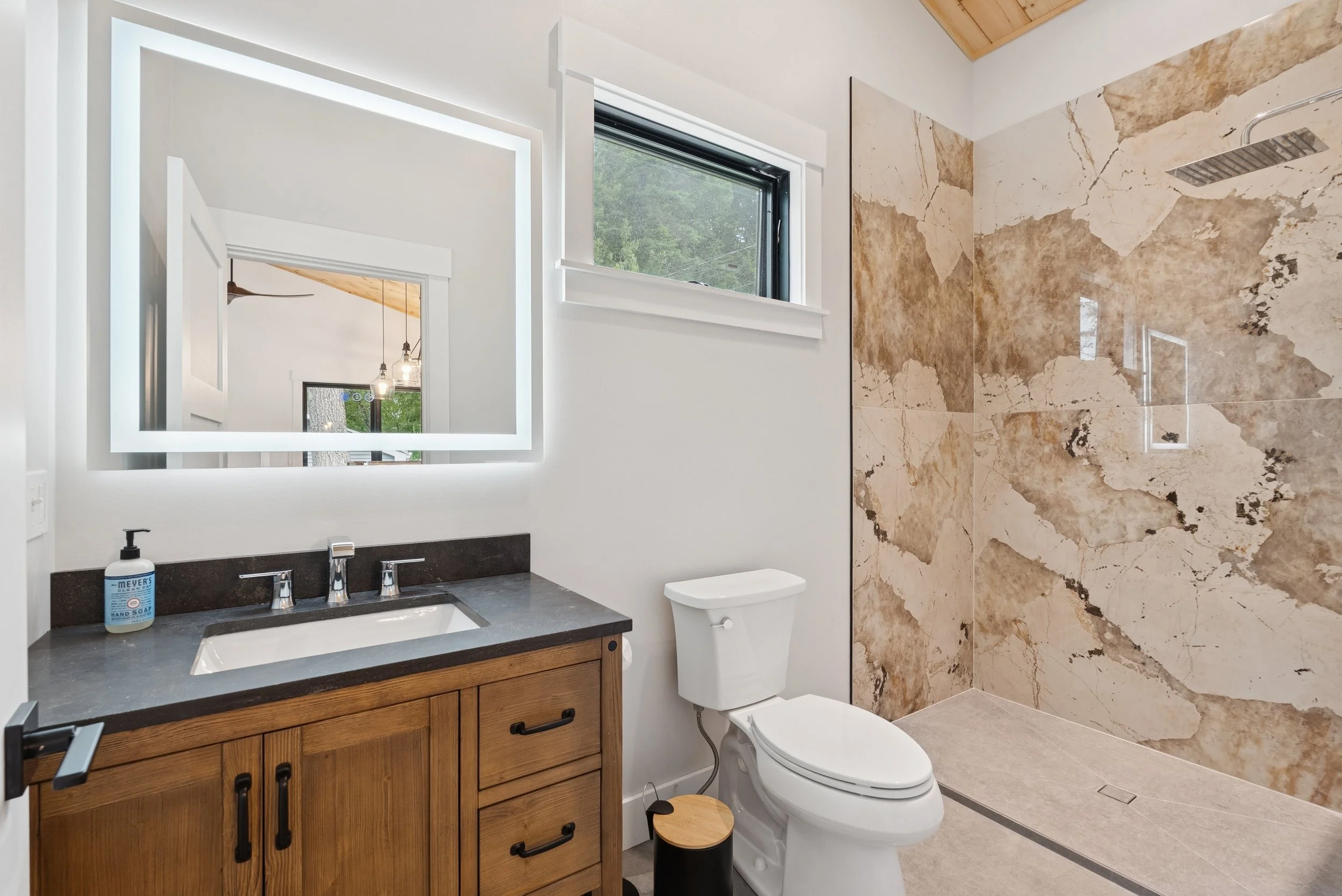 Modern bathroom with a large marble shower, white walls, a vanity with a dark countertop, a rectangular mirror with integrated lighting, a window, and a toilet.