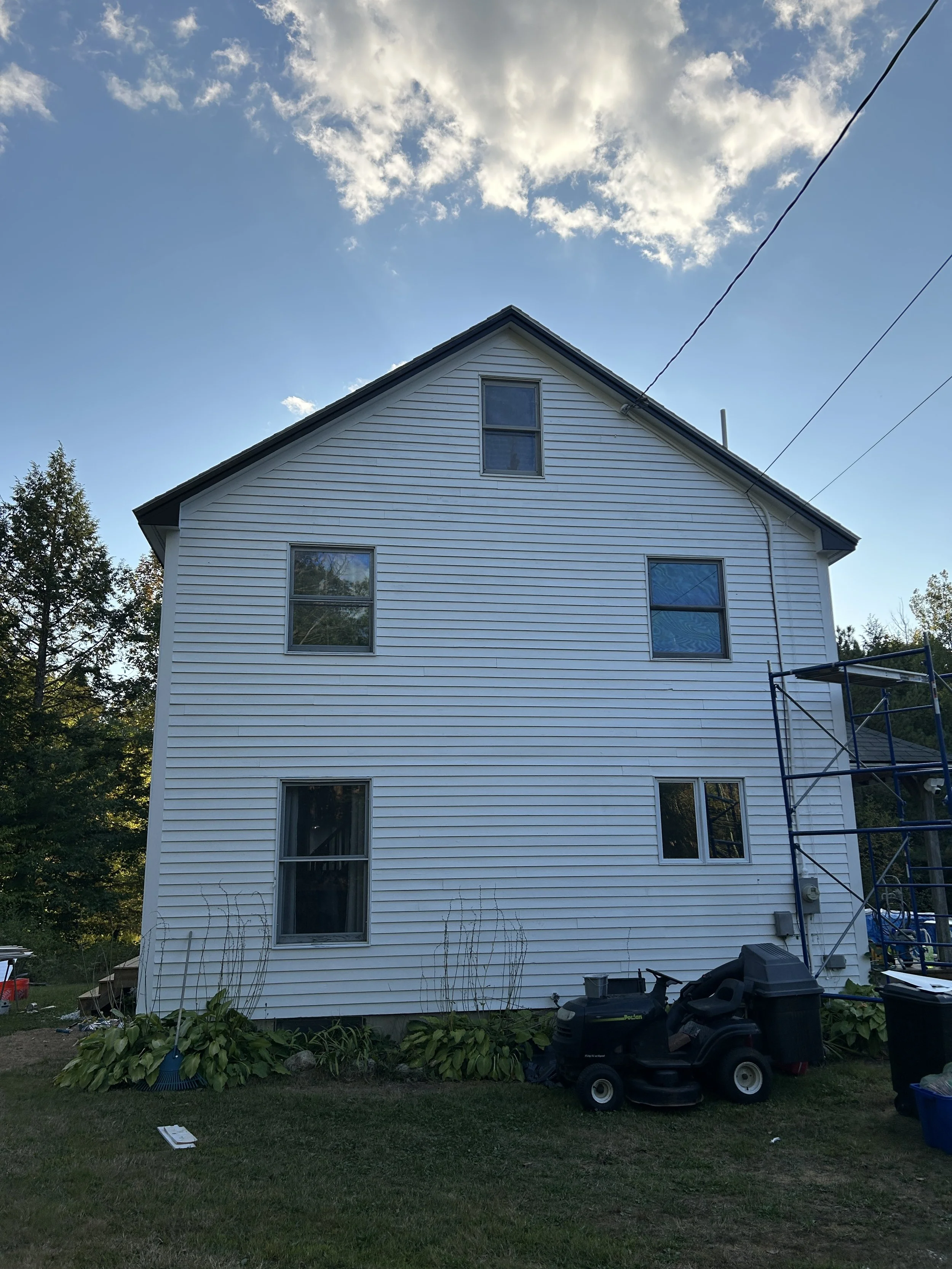 A white house with four visible windows, three on the main floor and one in the attic, with a gabled roof. There is scaffolding on the right side of the house, and gardening tools and equipment, including a lawnmower, are in the yard in front. The sk