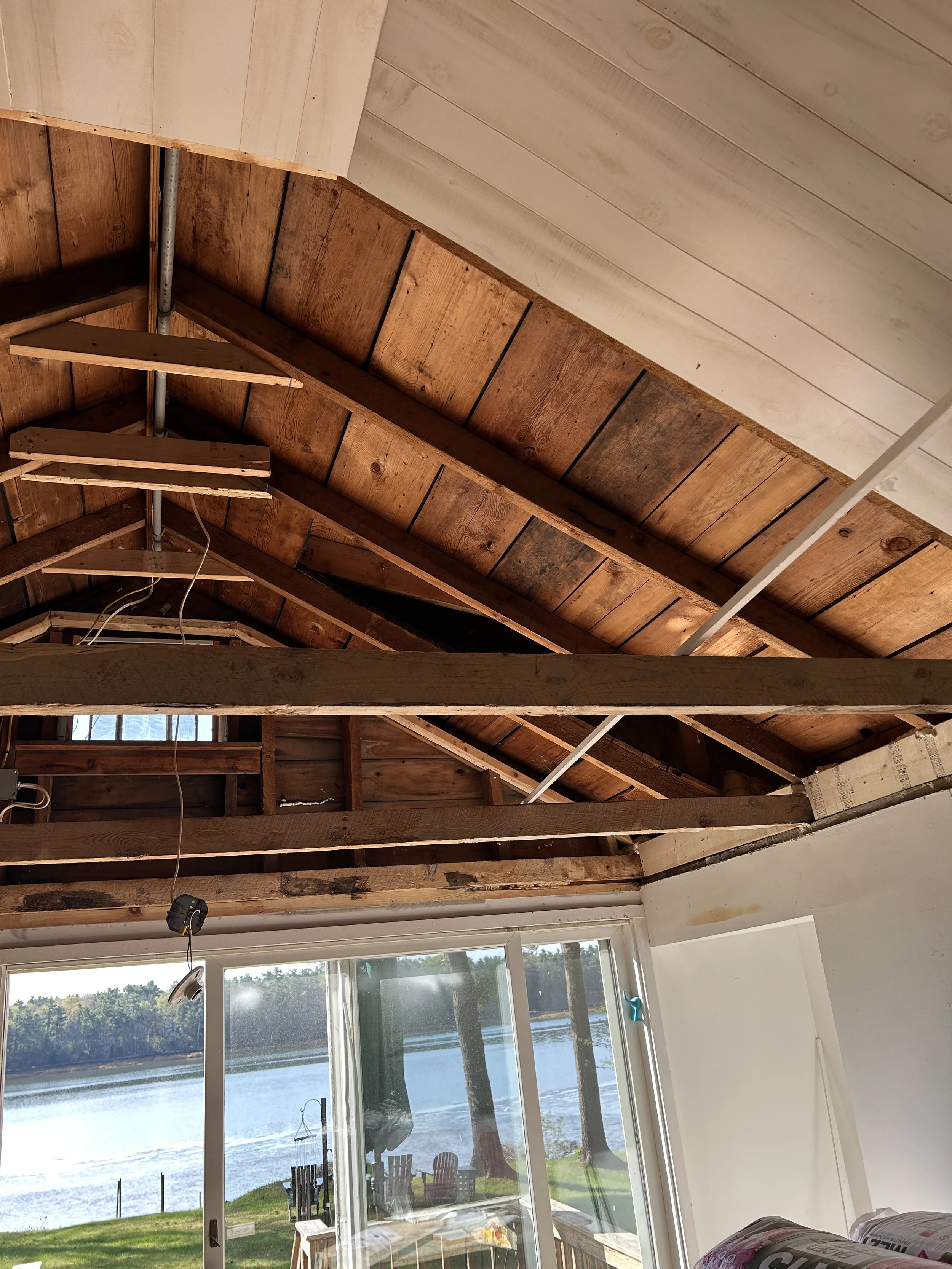 Interior view of a house undergoing construction or renovation, showing exposed wooden ceiling beams, a sliding glass door, and a view of a lake with trees and outdoor chairs