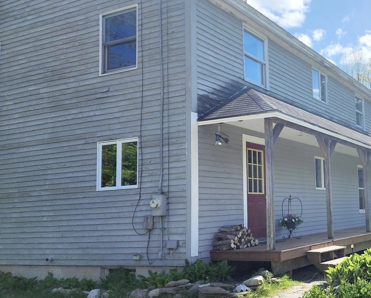 Exterior of a two-story house with gray siding, a small porch with stairs, a door, and several windows. There are logs stacked near the door and a plant in a decorative pot on the porch.