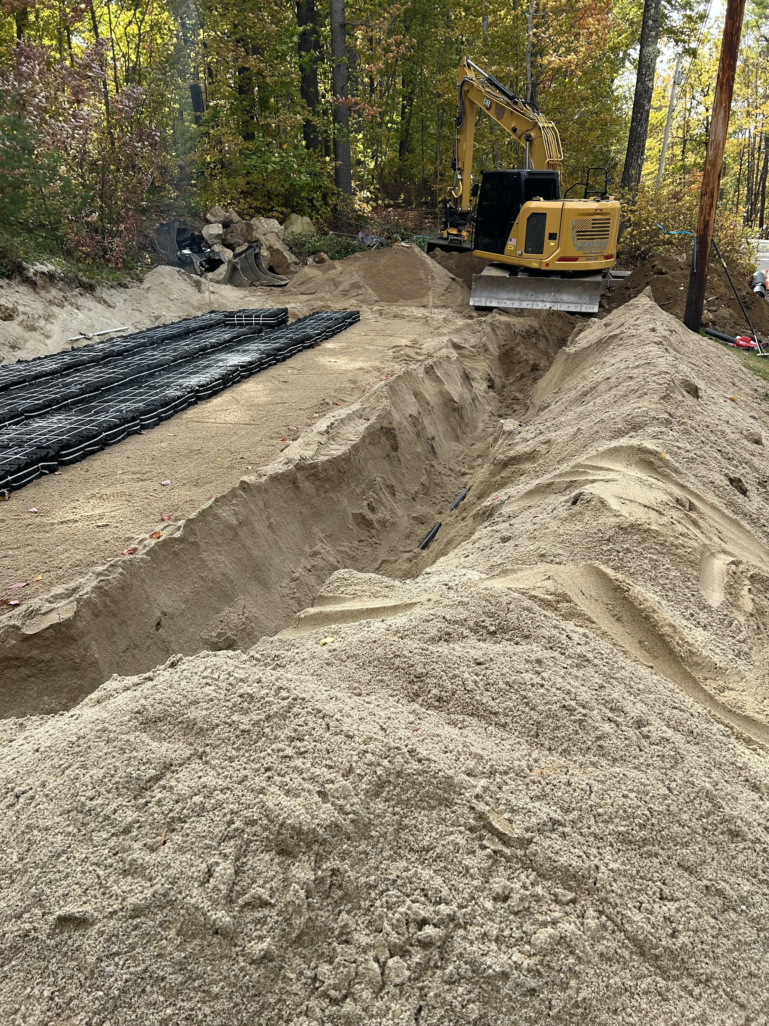 Construction site with a small yellow excavator working on a dirt trench amidst trees, with sand, rocks, and construction materials nearby.