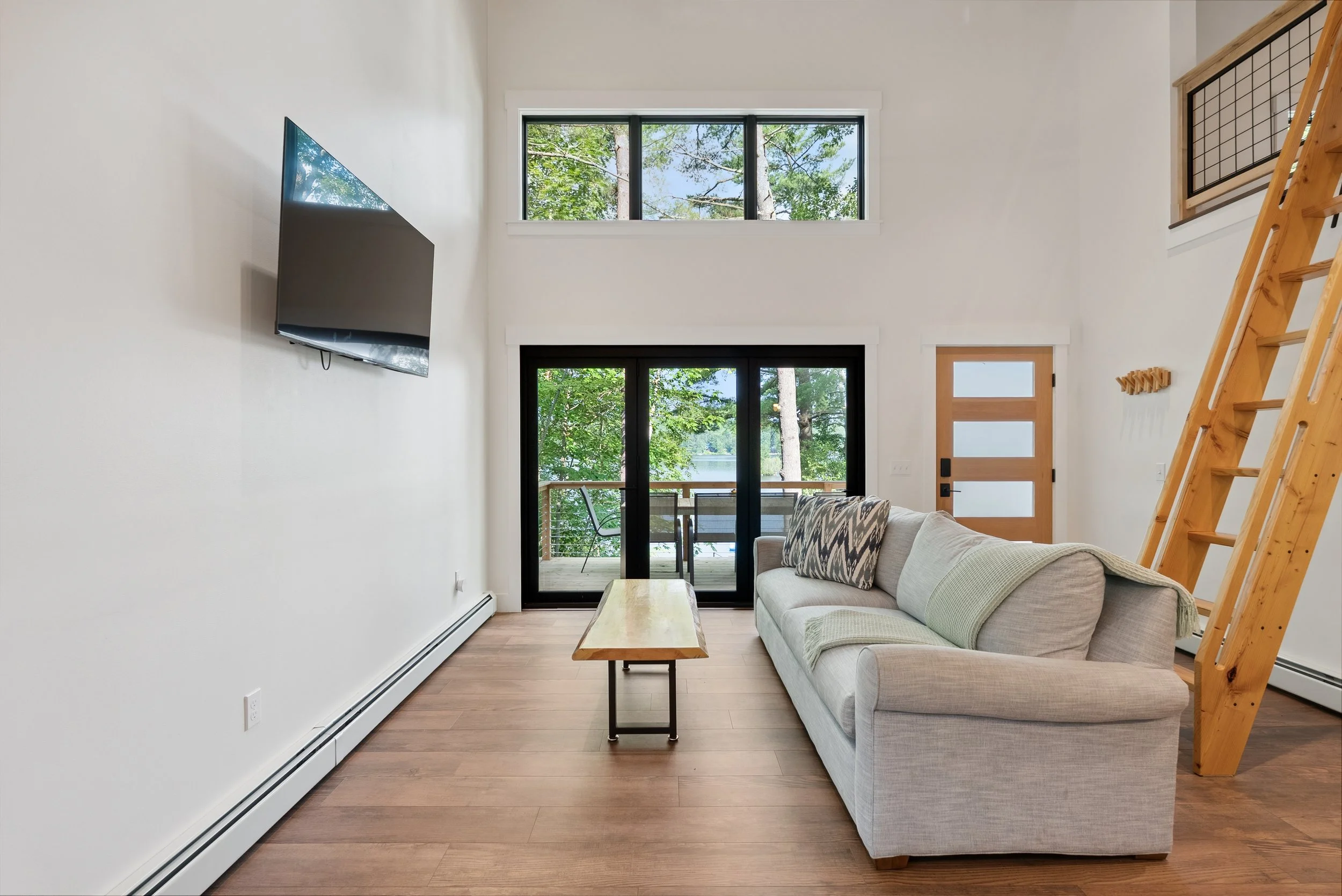 Modern living room with a light-colored sofa, wooden coffee table, large windows showing trees outside, a wooden staircase, and a sliding glass door opening to a balcony with outdoor seating.