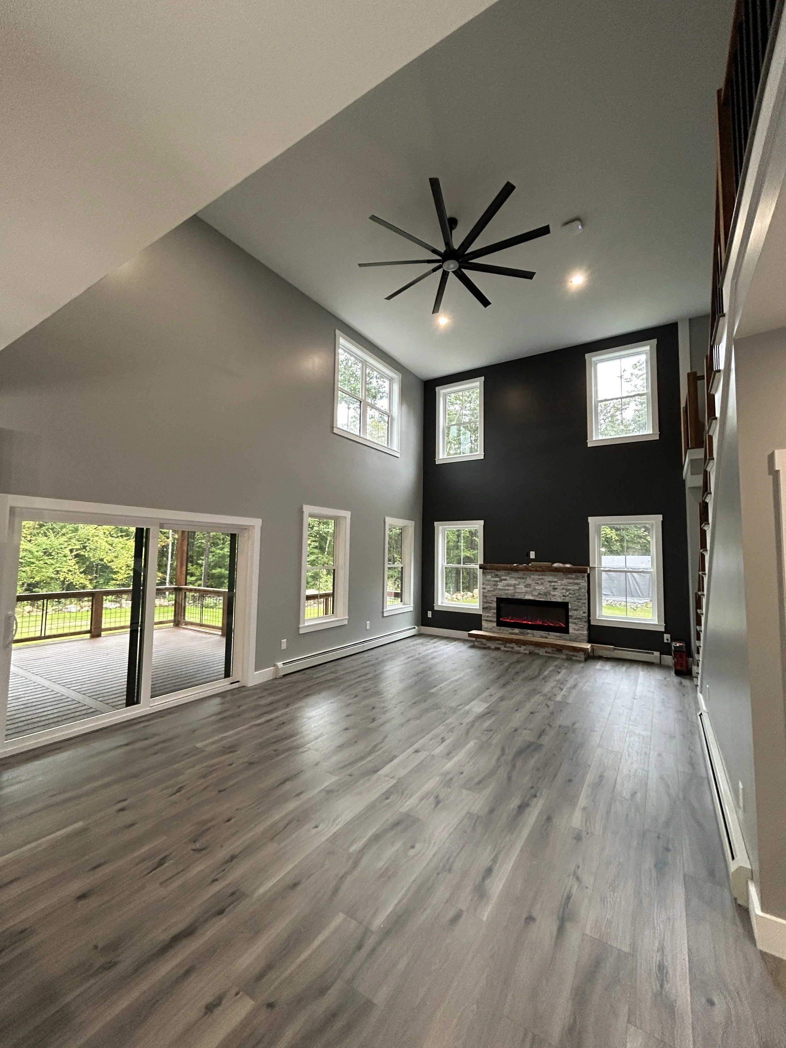 Empty living room with high ceiling, large windows, a fireplace, and wood flooring, with a balcony outside.