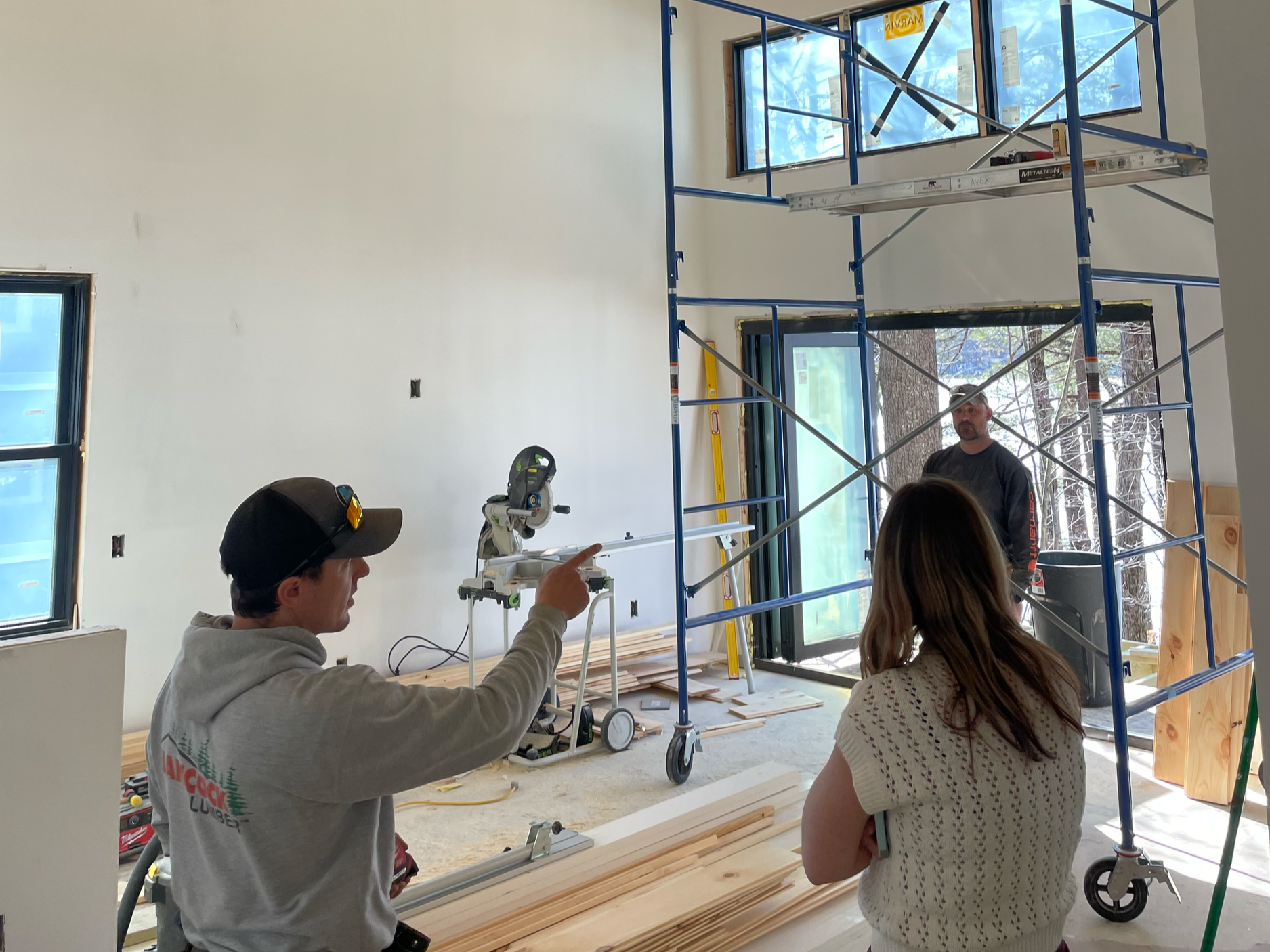 Construction workers inside a building under renovation, with scaffolding, woodworking tools, and large windows showing trees outside.