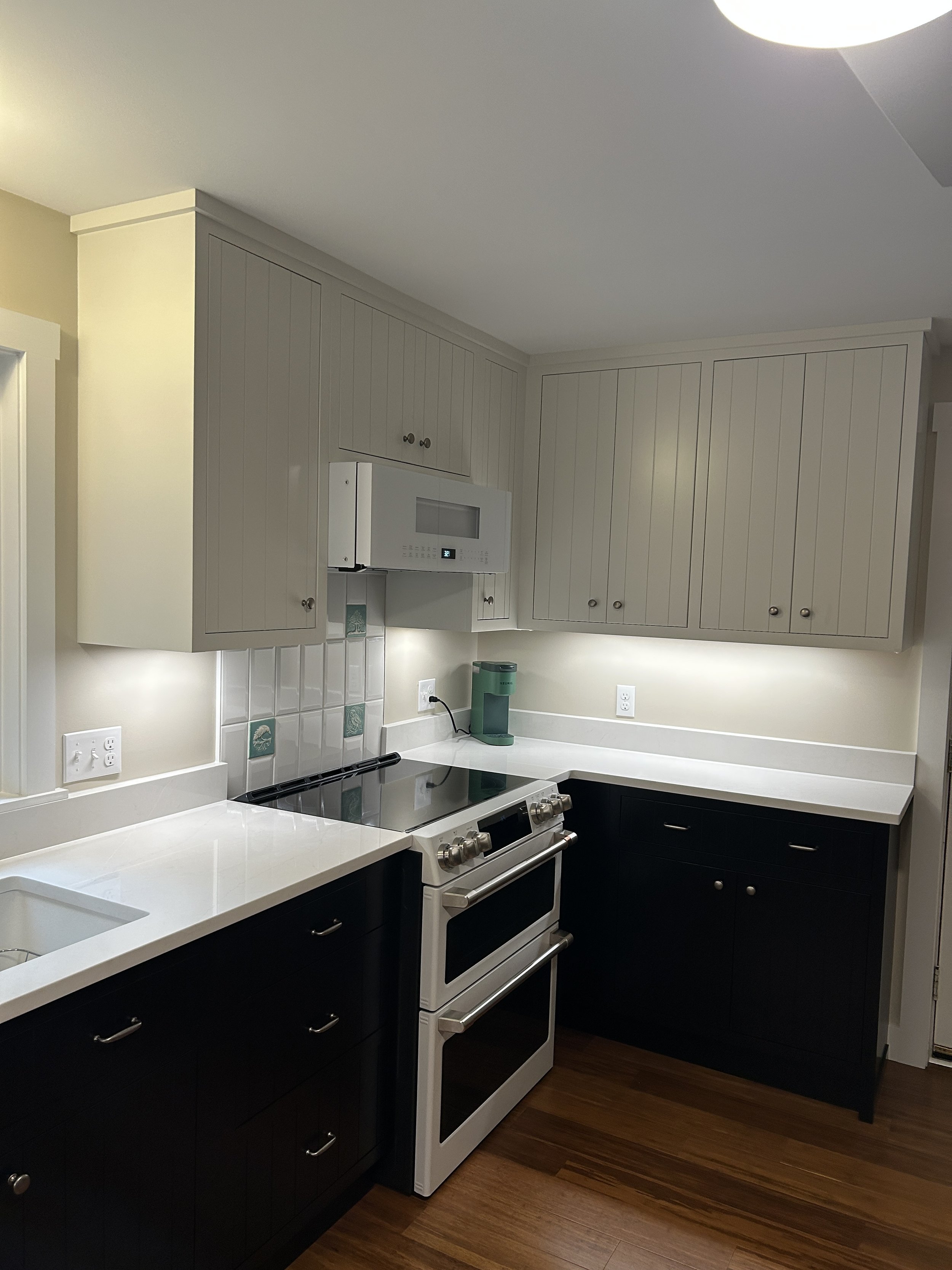 Kitchen with white and black cabinets, white countertops, a stove, and a microwave, with wooden flooring.