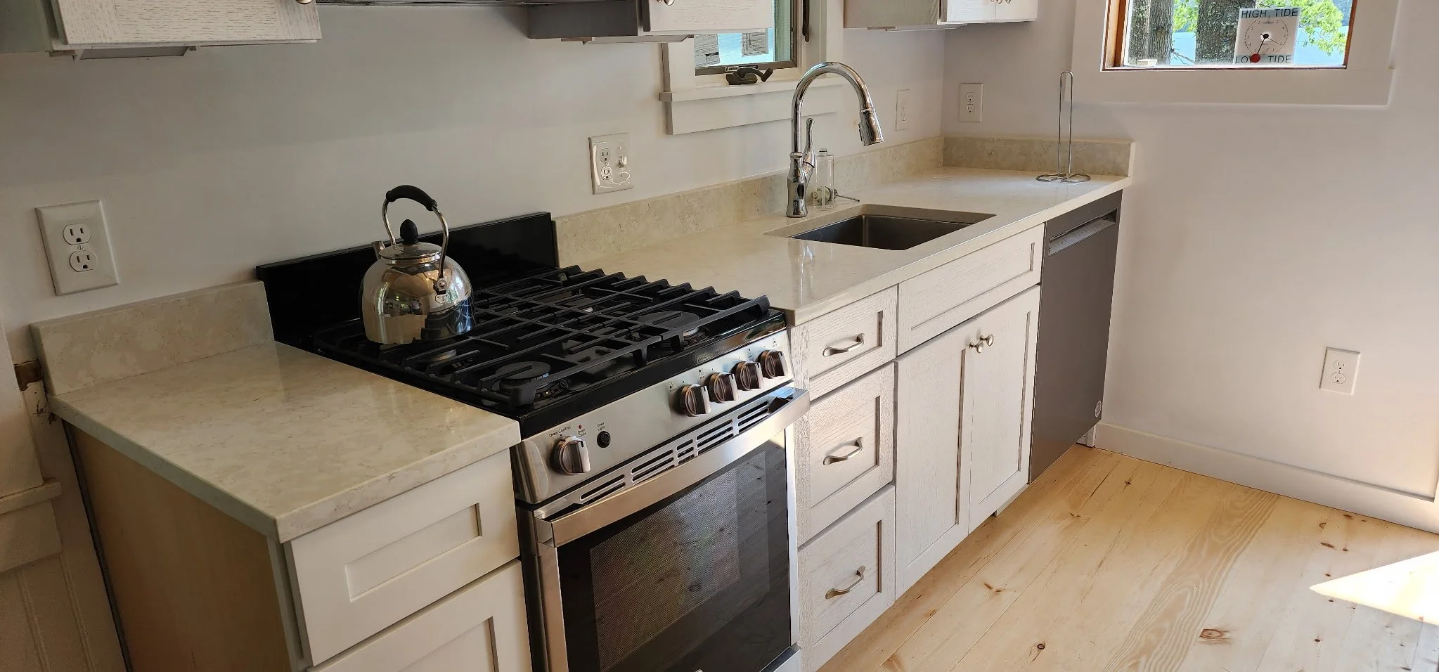 Kitchen with white cabinets, a beige countertop, a stainless steel stove with a kettle, a sink with a chrome faucet, and a small window with a high tide water level sign outside.