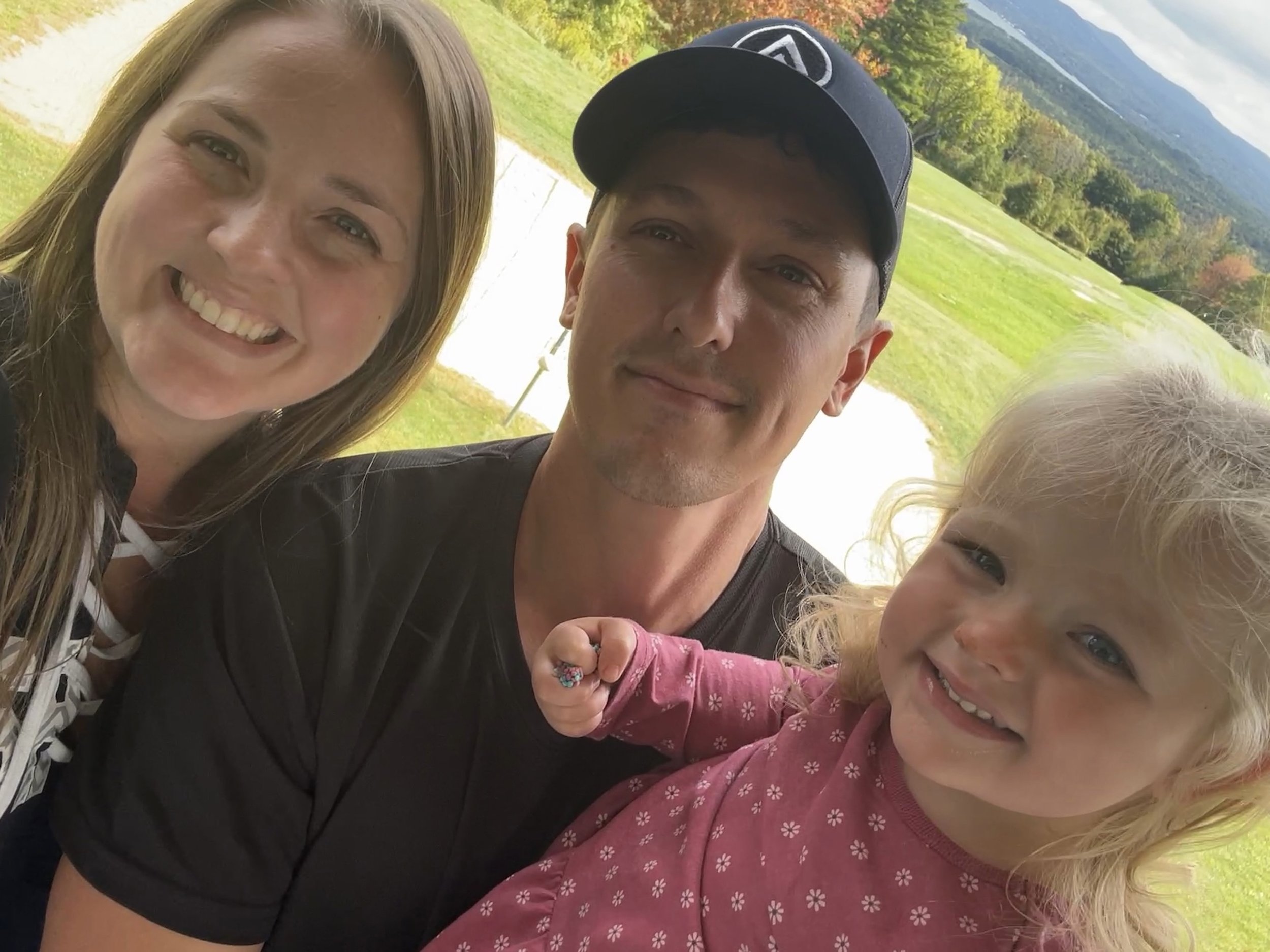 A group of smiling people, including a woman with light brown hair, a man wearing a black cap, and two young children with blonde hair, outdoors in a green, mountainous area.