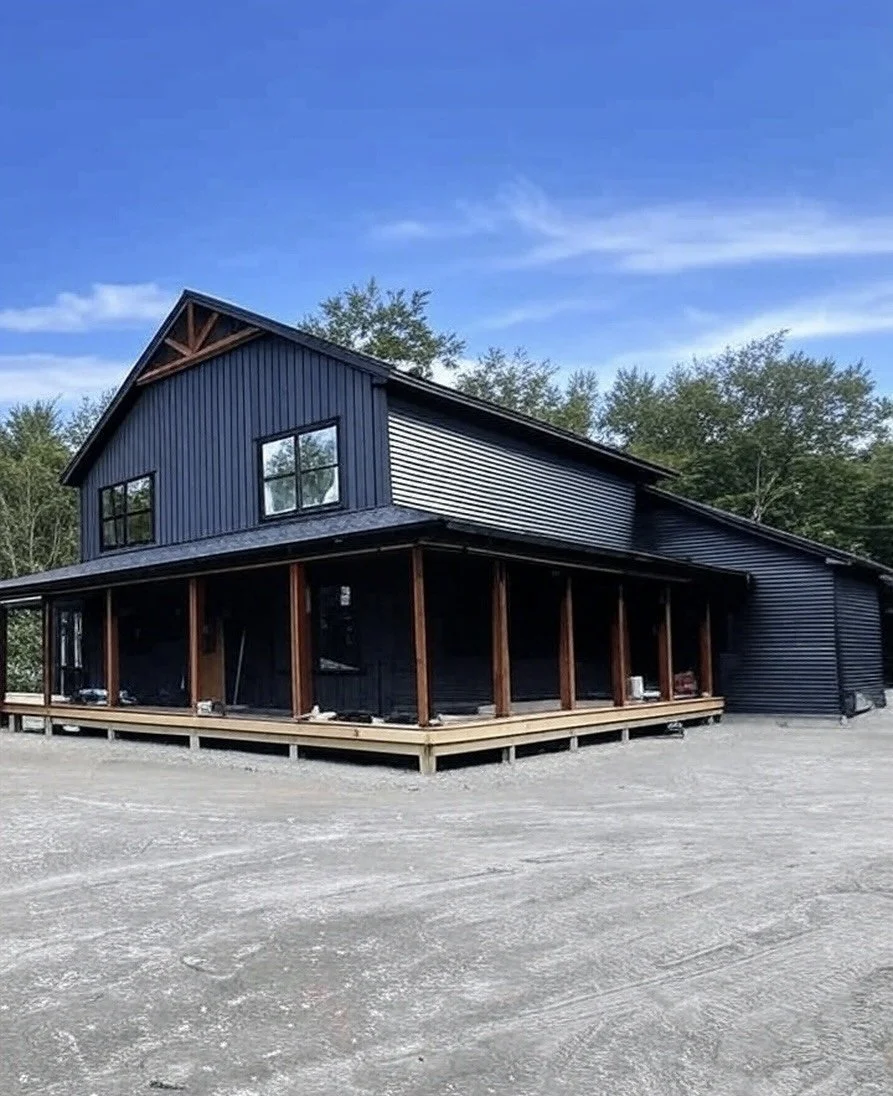 A two-story house with black siding and a covered porch under construction, surrounded by trees and a gravel driveway.