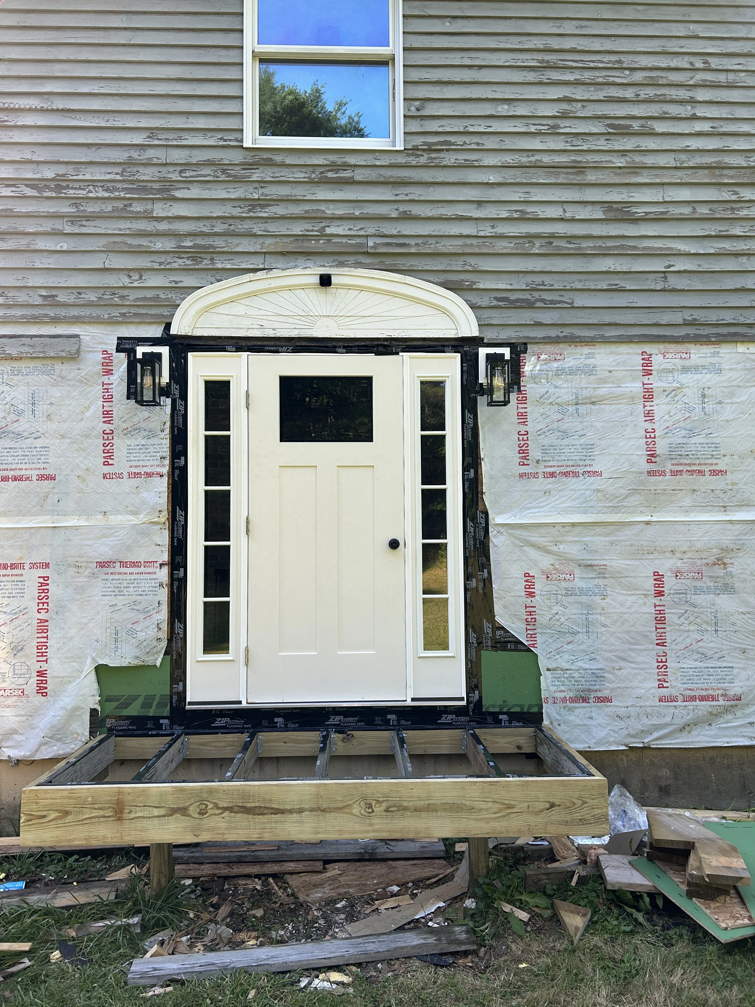 Construction site featuring a front door with sidelight windows, partially wrapped in black tape, with a wooden frame below for porch construction, and siding being installed on the house exterior with exposed sheathing and insulation.