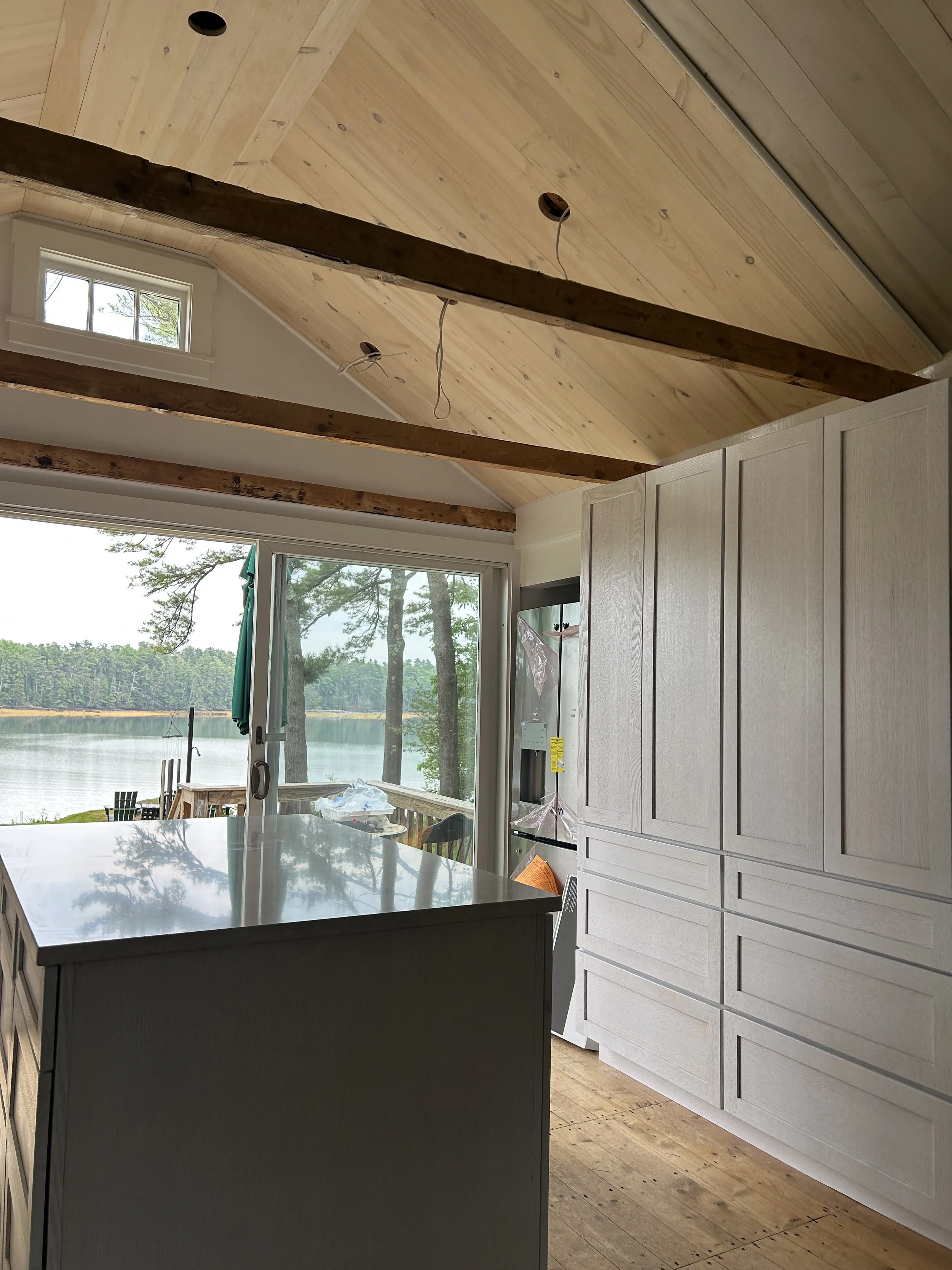 A kitchen with a view of a lake and trees outside large glass doors, with white cabinets, a dark gray island, and a light wooden sloped ceiling with exposed beams.