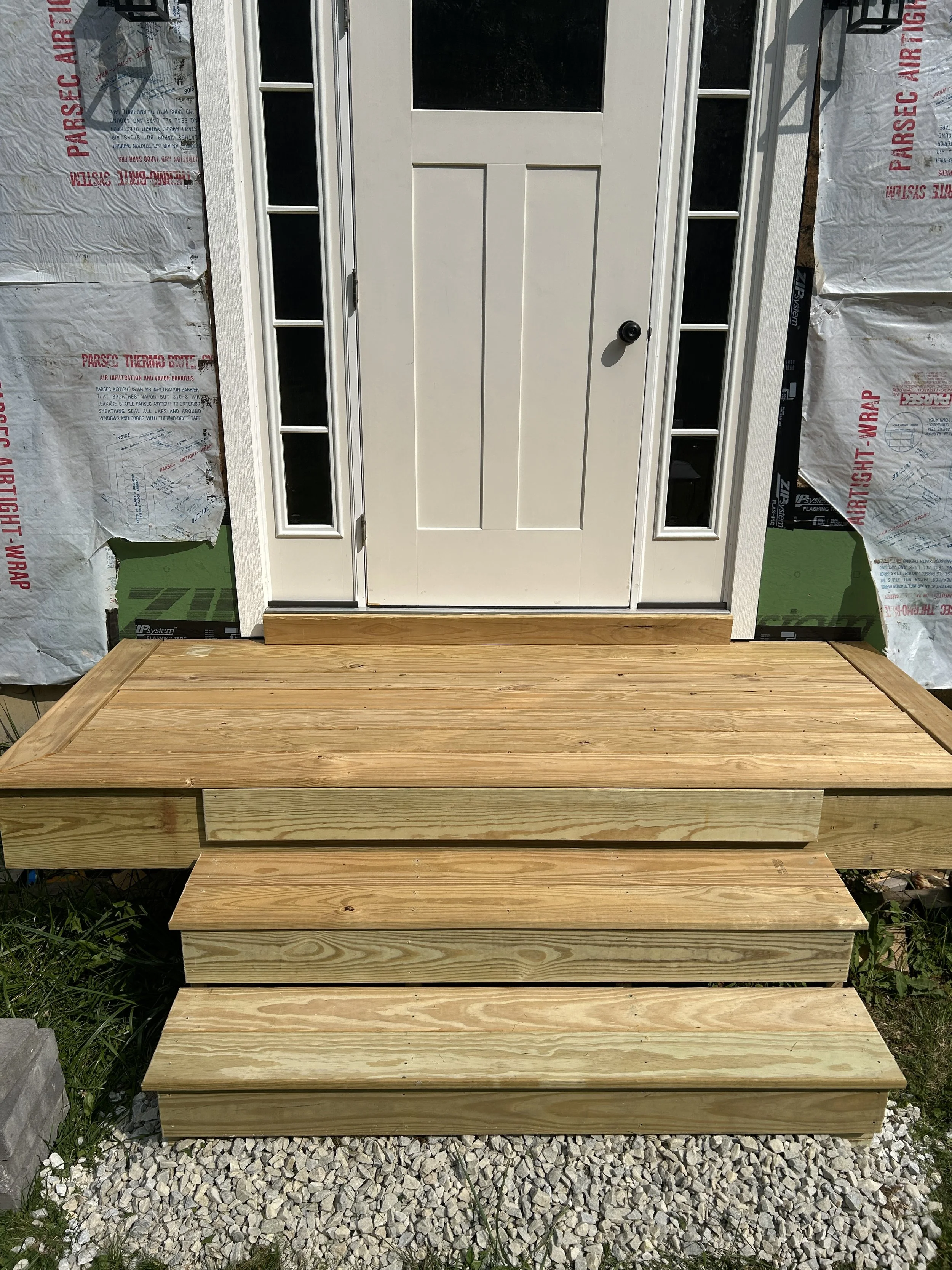 A white front door with a small window at the top and a black doorknob, surrounded by white framed side windows, with a wooden porch and steps leading up to it, under construction with insulation material on the sides.