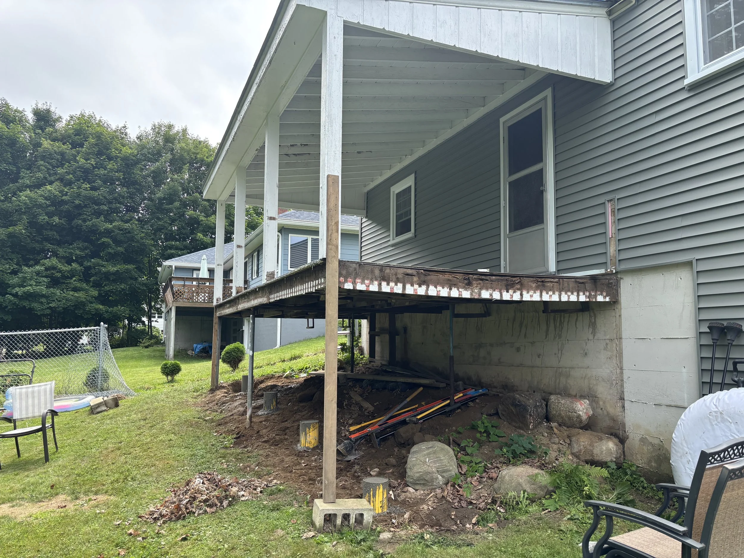The image shows the backyard of a house with a deck under construction. The house has gray siding and several windows. There are construction tools and materials, including wooden columns, rocks, and digging equipment, indicating ongoing remodeling w