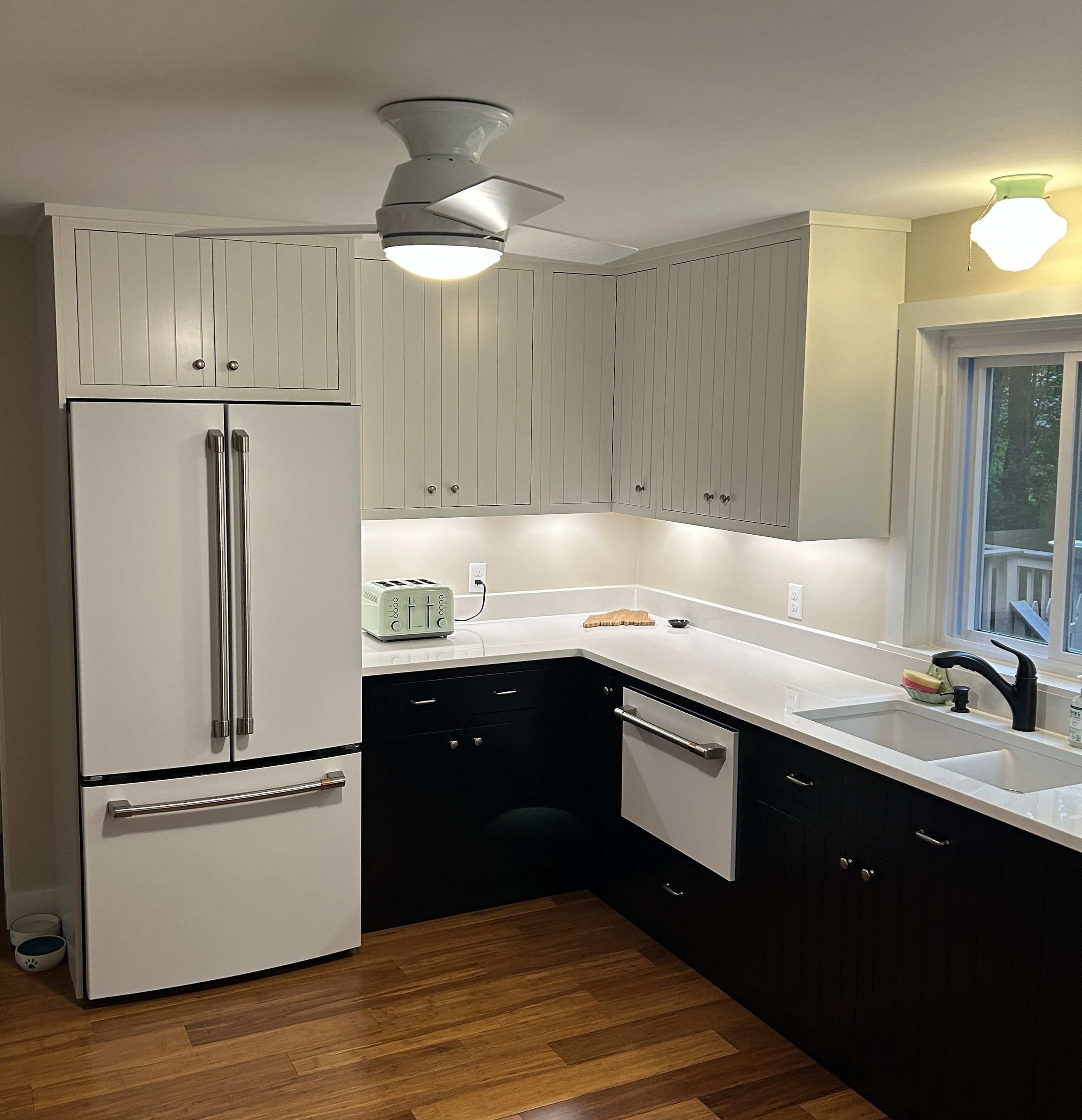 View of a kitchen corner with white upper cabinets, black lower cabinets, a white countertop, a white refrigerator, a pale yellow wall, a window with outside trees, and a ceiling fan with a light. There's also a toaster, a soap dispenser, and a dog f