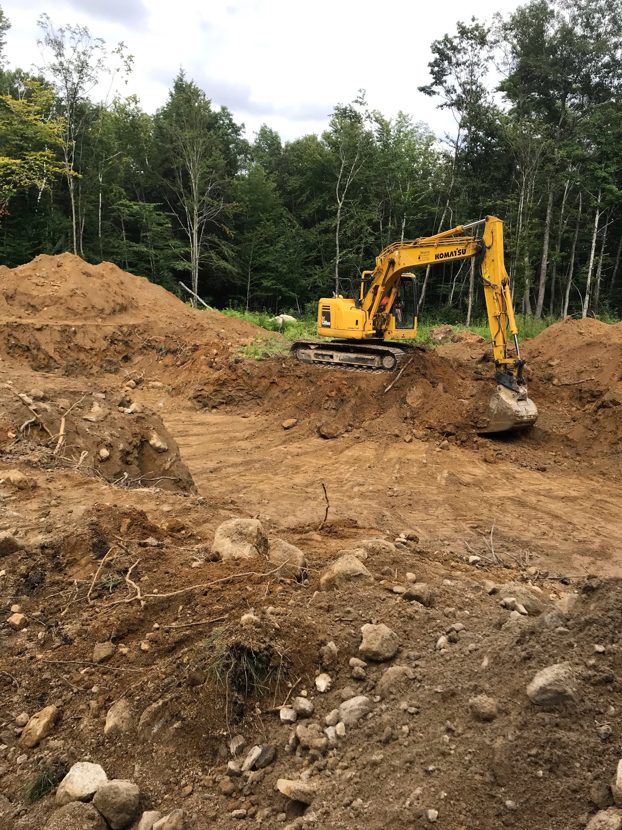 A small yellow excavator working on a dirt construction site surrounded by rocks, with a forest of tall trees in the background.