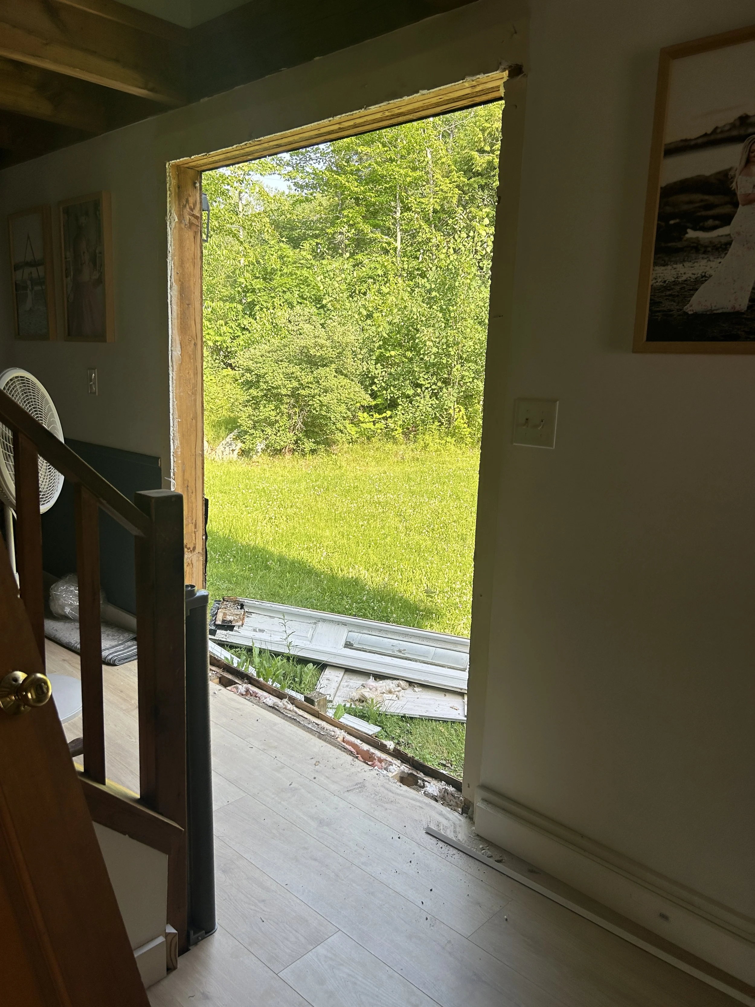 View of a house door missing, revealing a lush green outdoor landscape with trees and grass. Inside, a staircase and framed pictures on the wall are visible, with construction debris near the threshold.