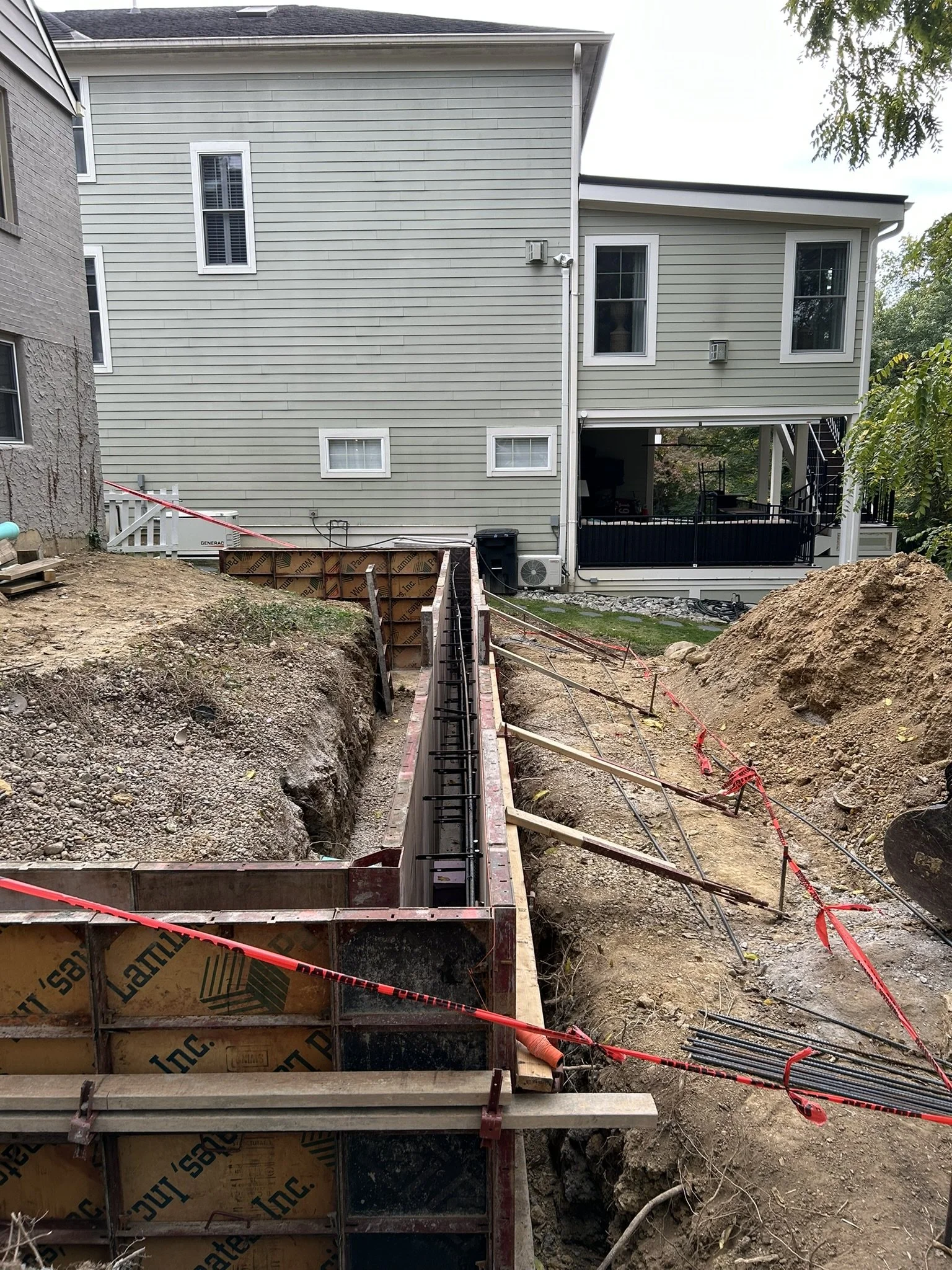 Construction site with exposed foundation and rebar, dug-out earth, and a partially constructed wall with plywood sheathing. The background shows a house with beige siding and multiple windows.