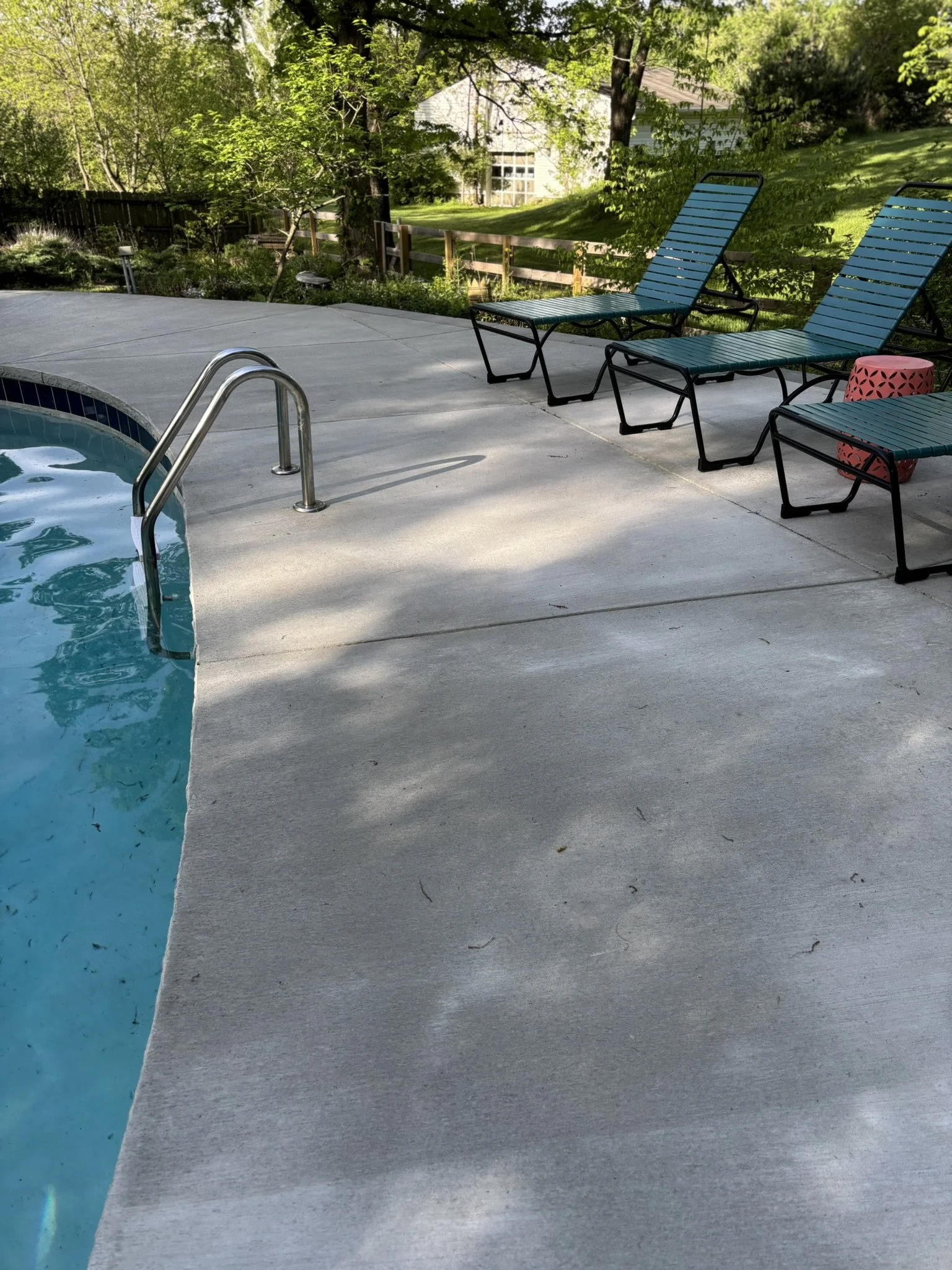 A backyard with a swimming pool, blue lounge chairs, a pink stool, trees, and a house in the background.