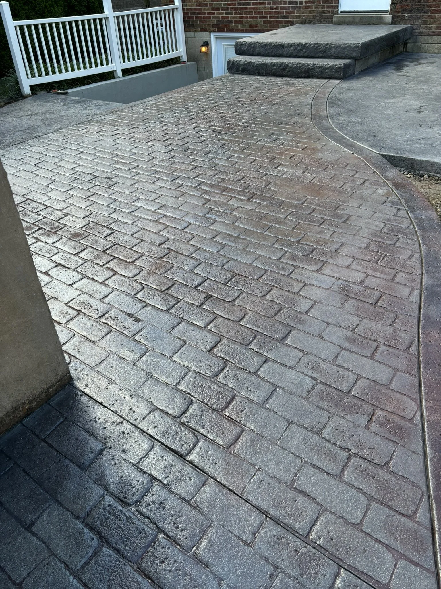 A newly paved walkway composed of small brick pavers, partially wet, with concrete steps leading to a door in a brick house. A white metal railing is on the left side of the walkway.