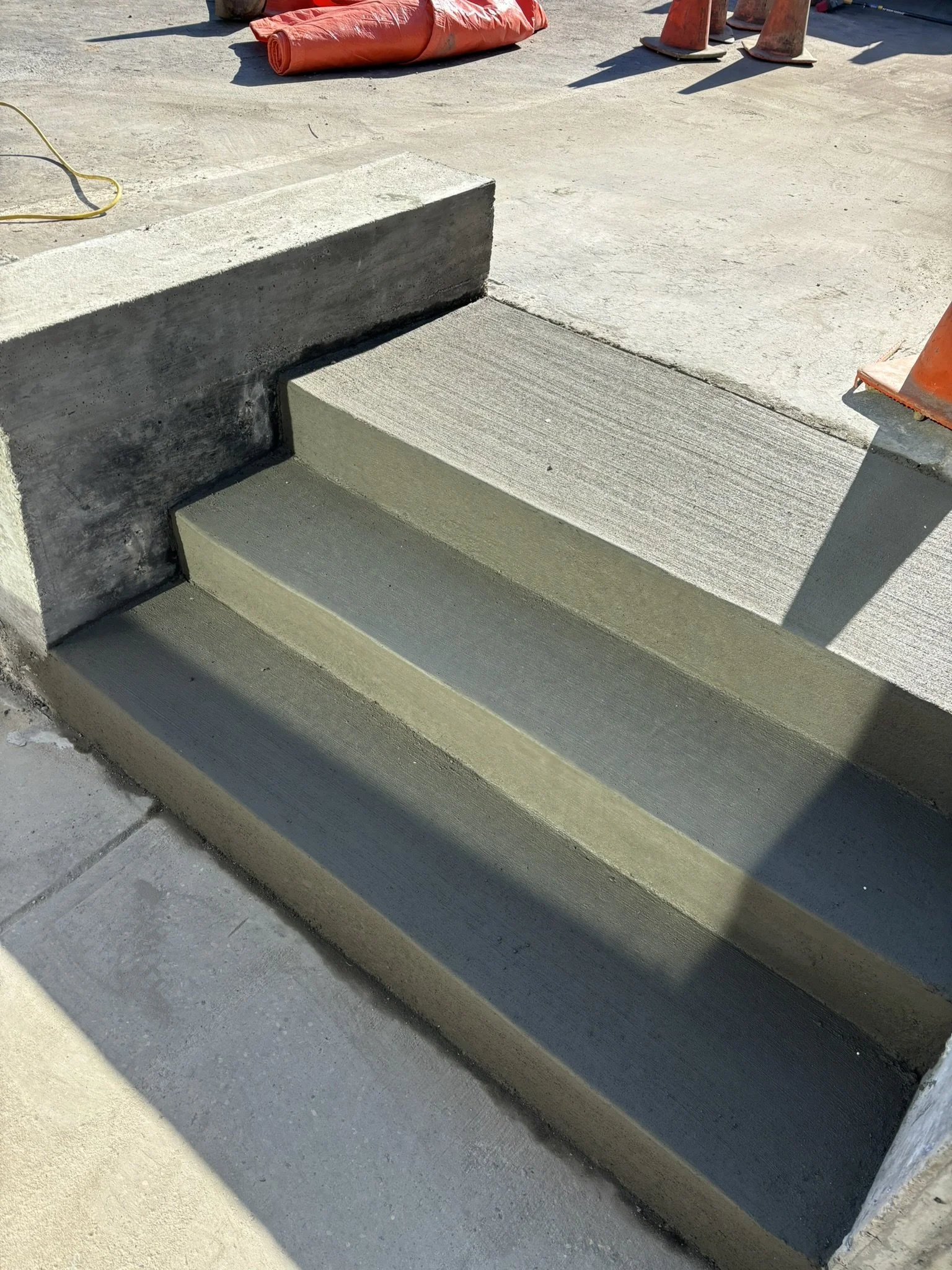 Construction site showing concrete stairs being built, orange construction barriers and a yellow extension cord on the ground.