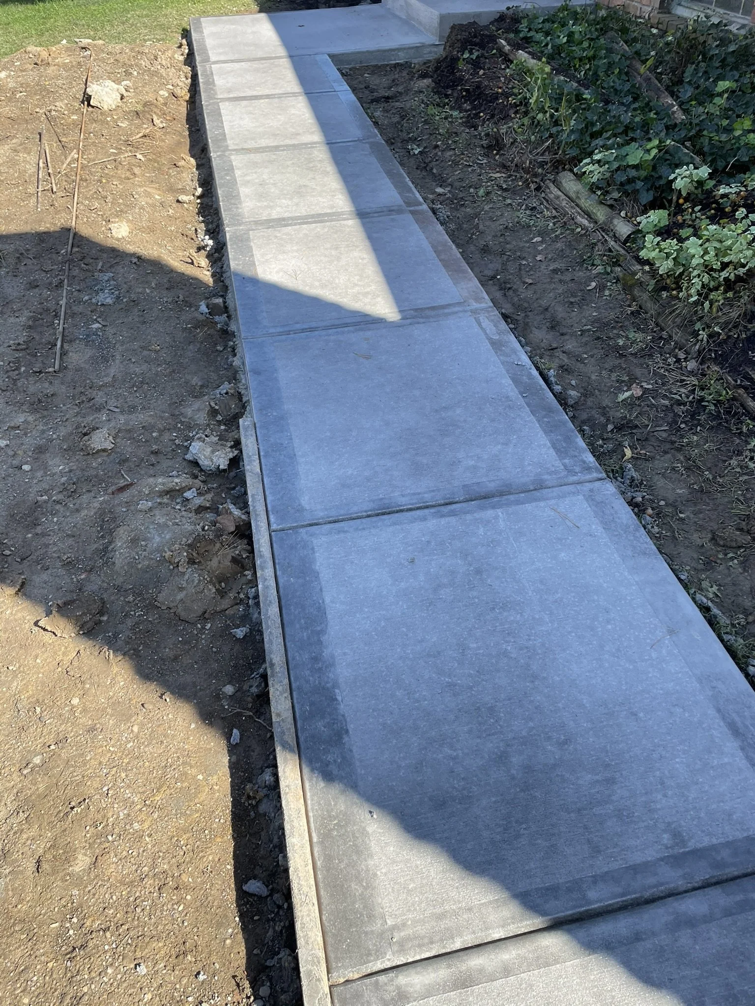New concrete sidewalk installed next to a garden bed with green plants, with dirt and debris on the adjacent side.