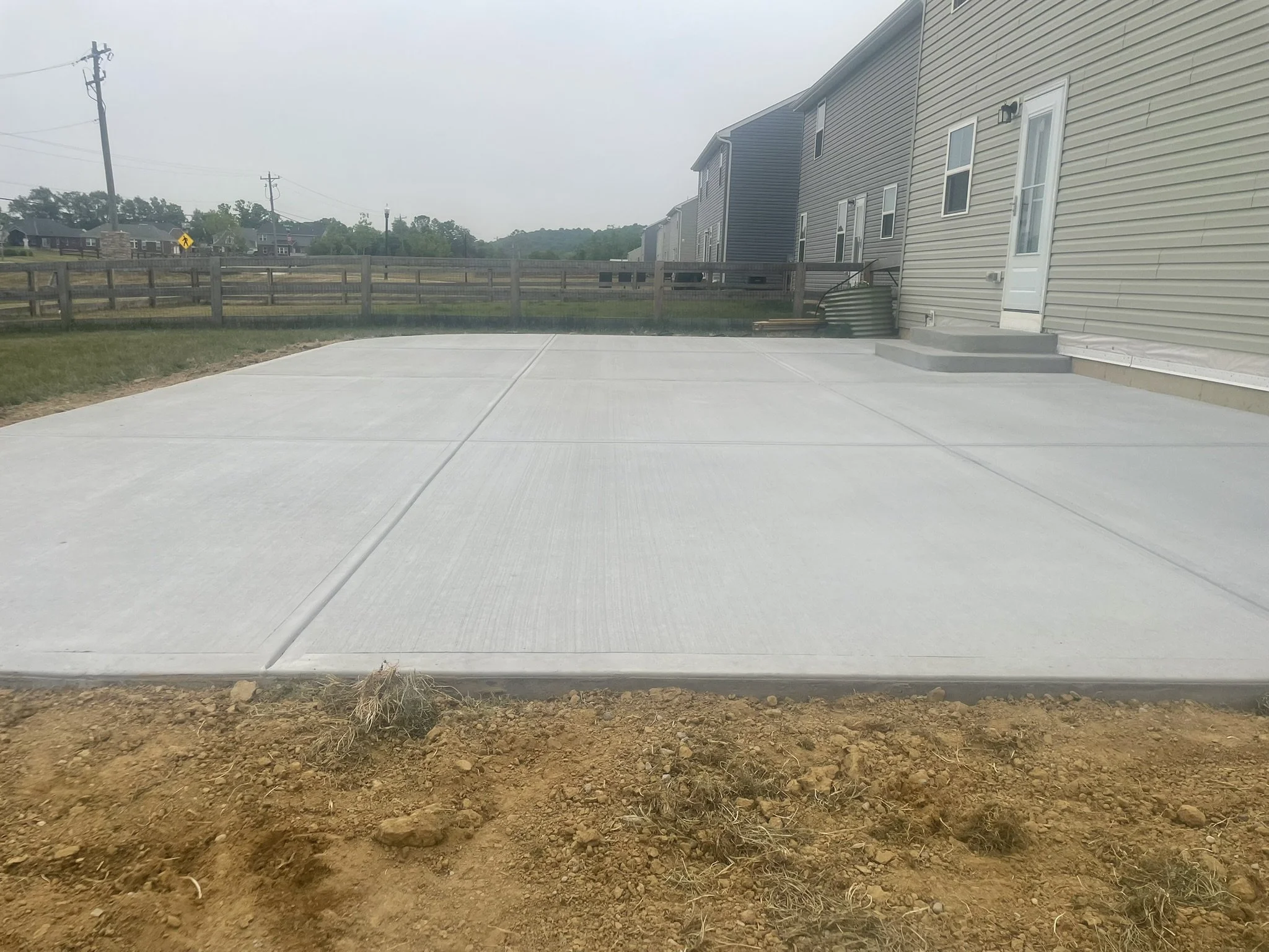 Newly poured concrete patio in backyard with surrounding dirt and grass, beige and blue houses in the background, and a wooden fence.
