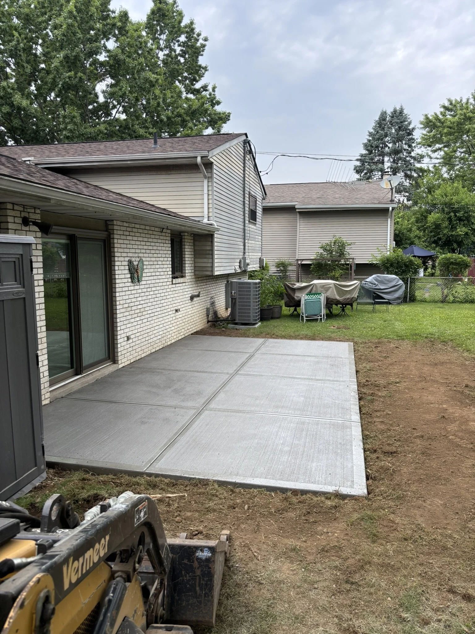 View of a backyard with a concrete patio, an air conditioning unit, and a table with chairs and covered grill, bordered by a lawn and a chain-link fence with green trees and neighboring houses in the background.